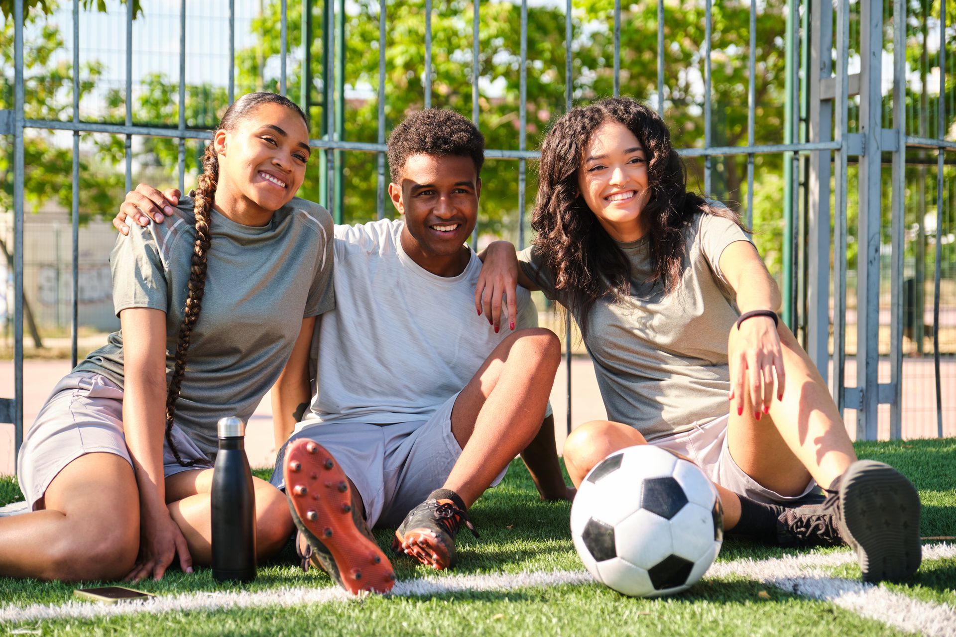 Three friends on a soccer field, smiling, two with arms around the other, soccer ball present.