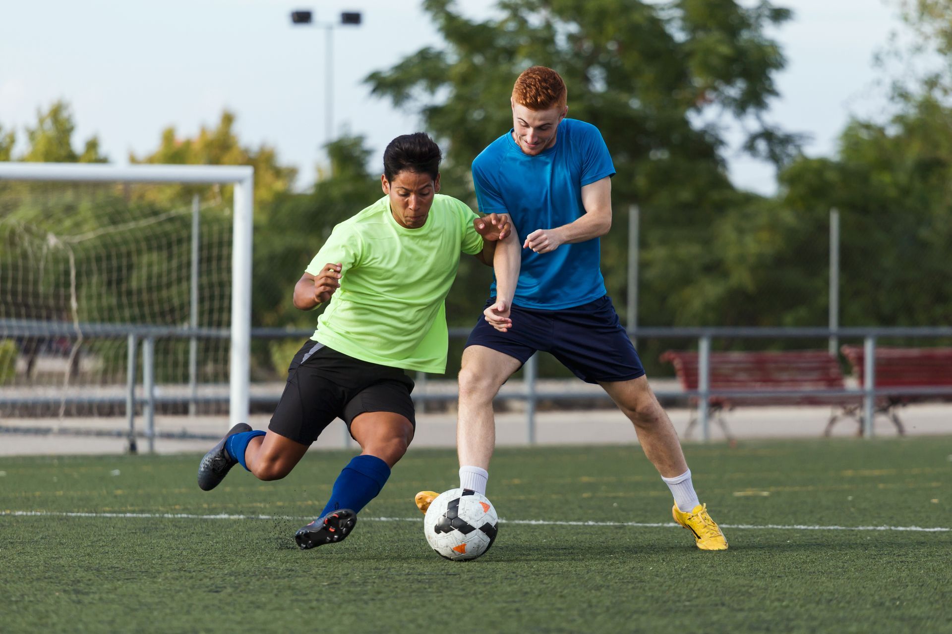 Two soccer players on a field: One in green shirt with ball, one in blue shirt reaching, near a goal.