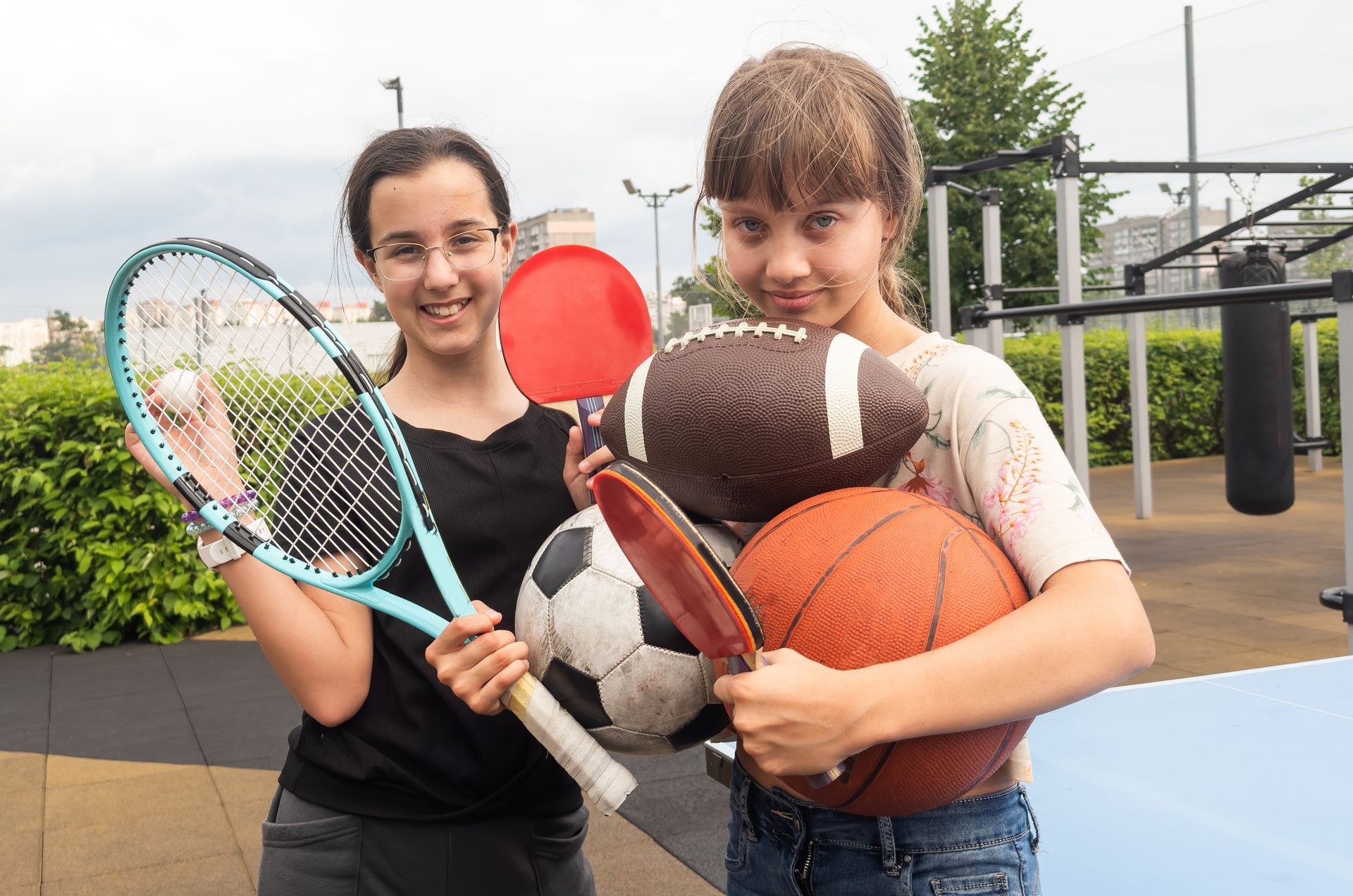 Two girls holding sports equipment: tennis racket, ping pong paddle, football, soccer ball, basketball, in an outdoor park.