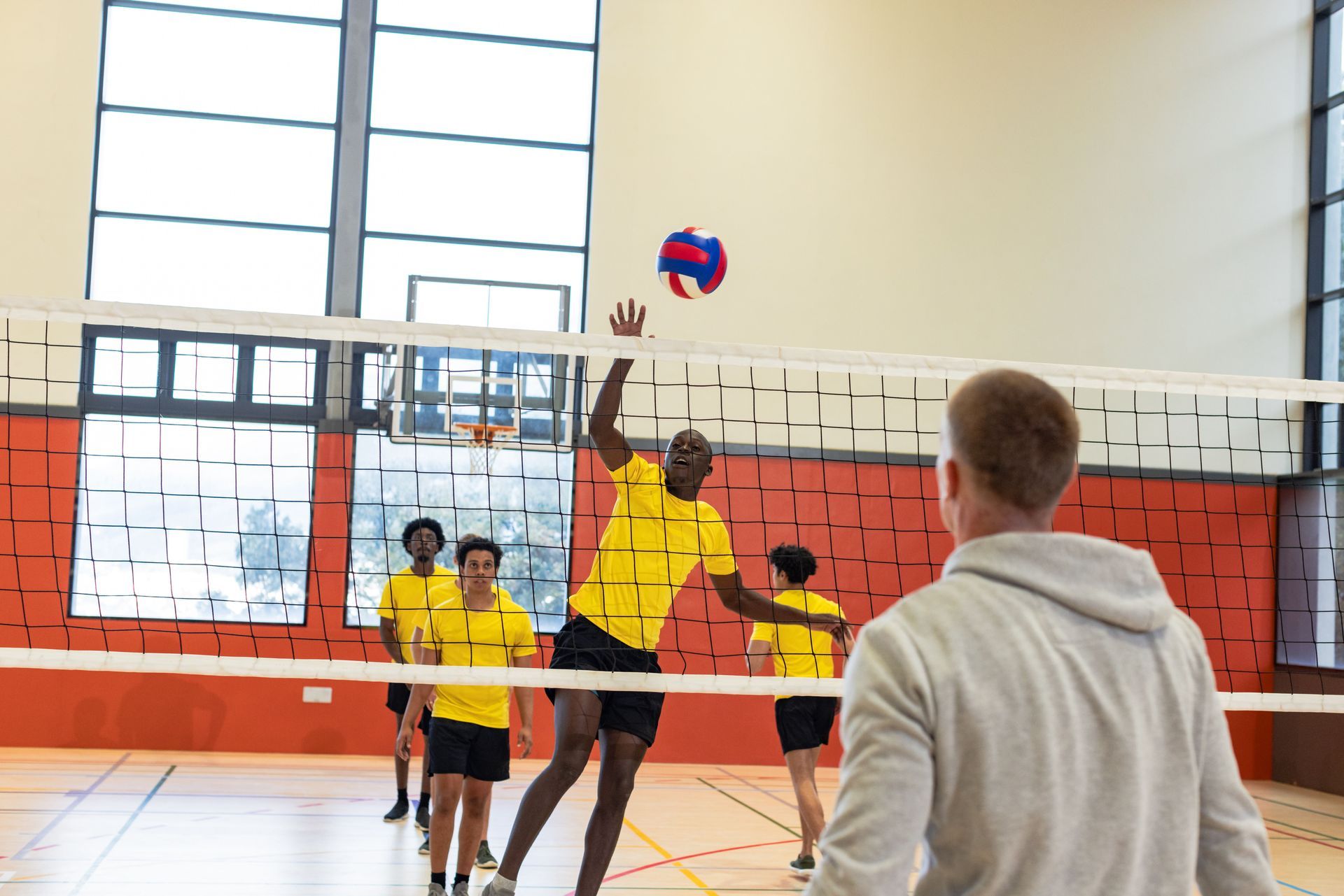 Volleyball player spiking the ball over the net in a gymnasium. Yellow uniforms, wooden floor.
