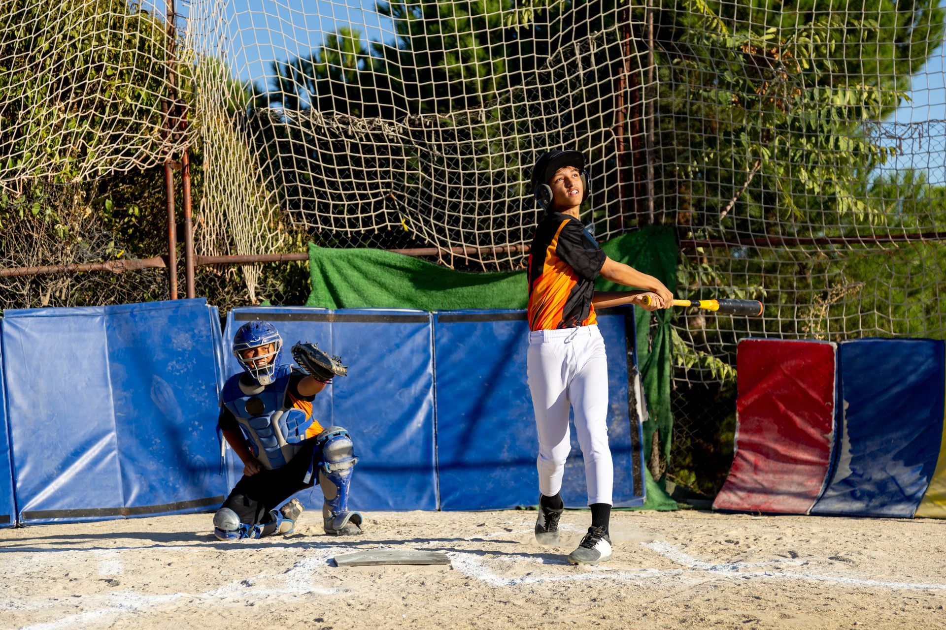 Batter swings at a ball in a baseball batting cage. Catcher crouches behind him.