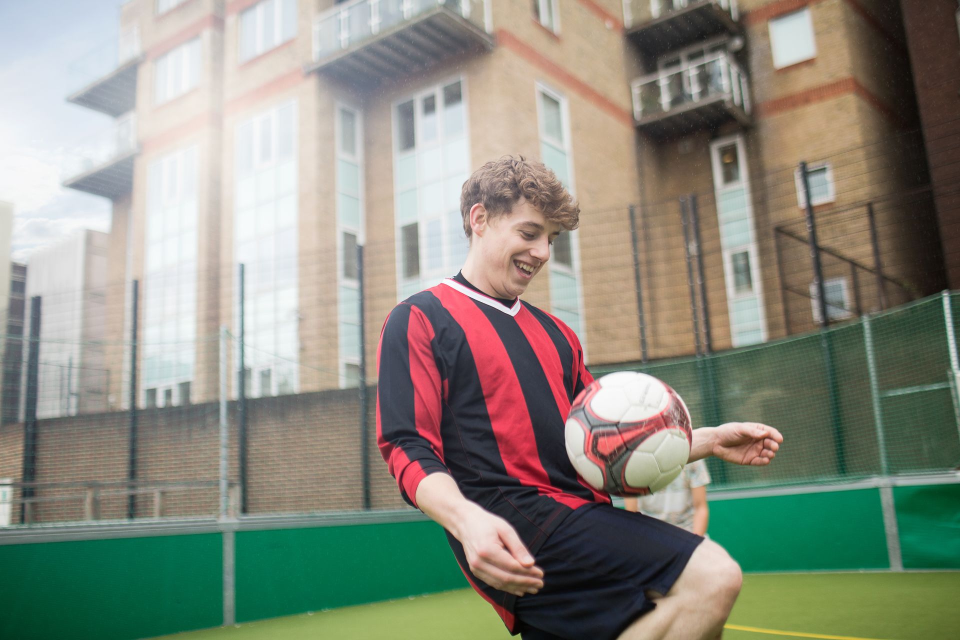 Soccer player in red/black jersey juggling ball outside a building.