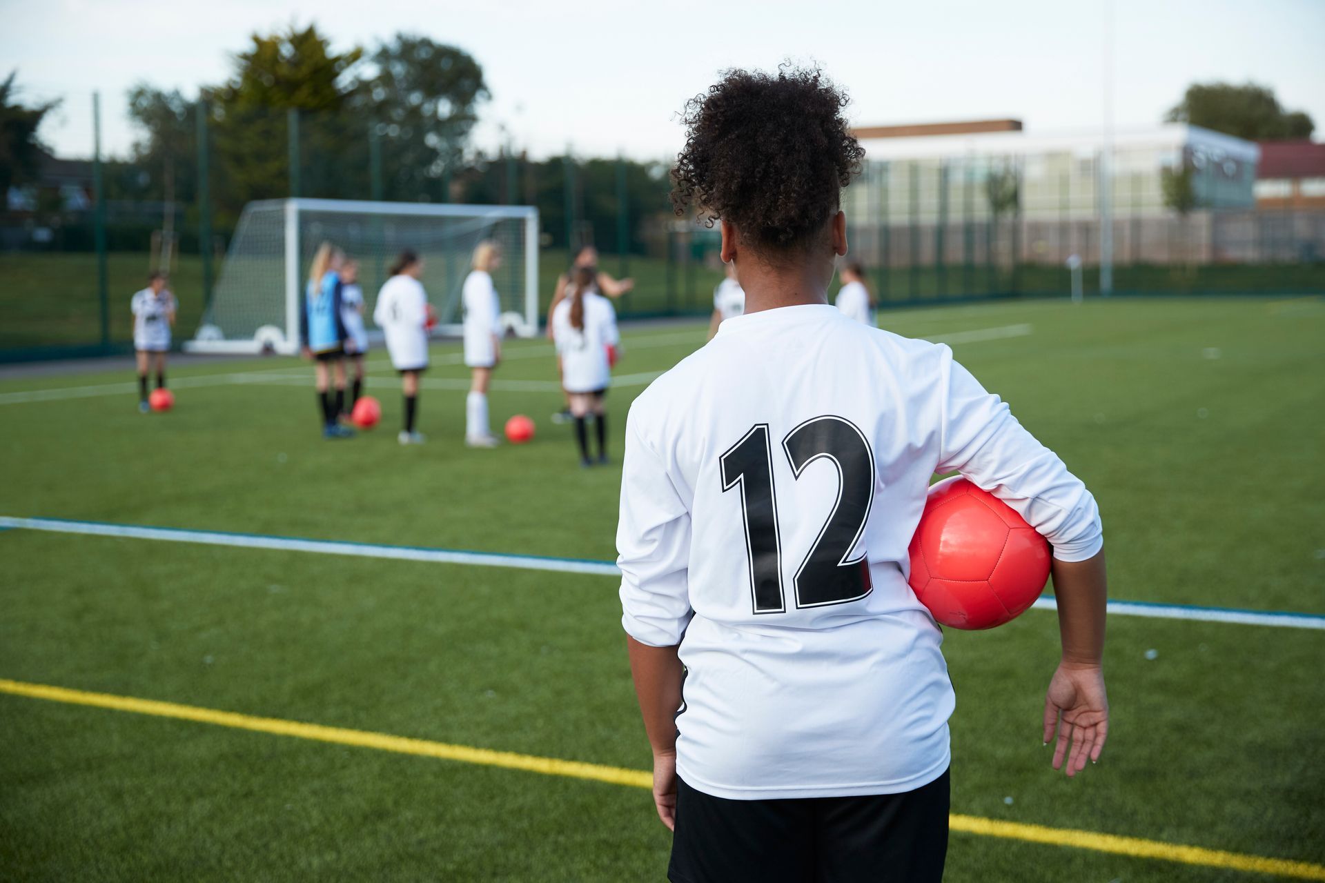 Soccer player, number 12, on field, holding ball, team practicing in background.