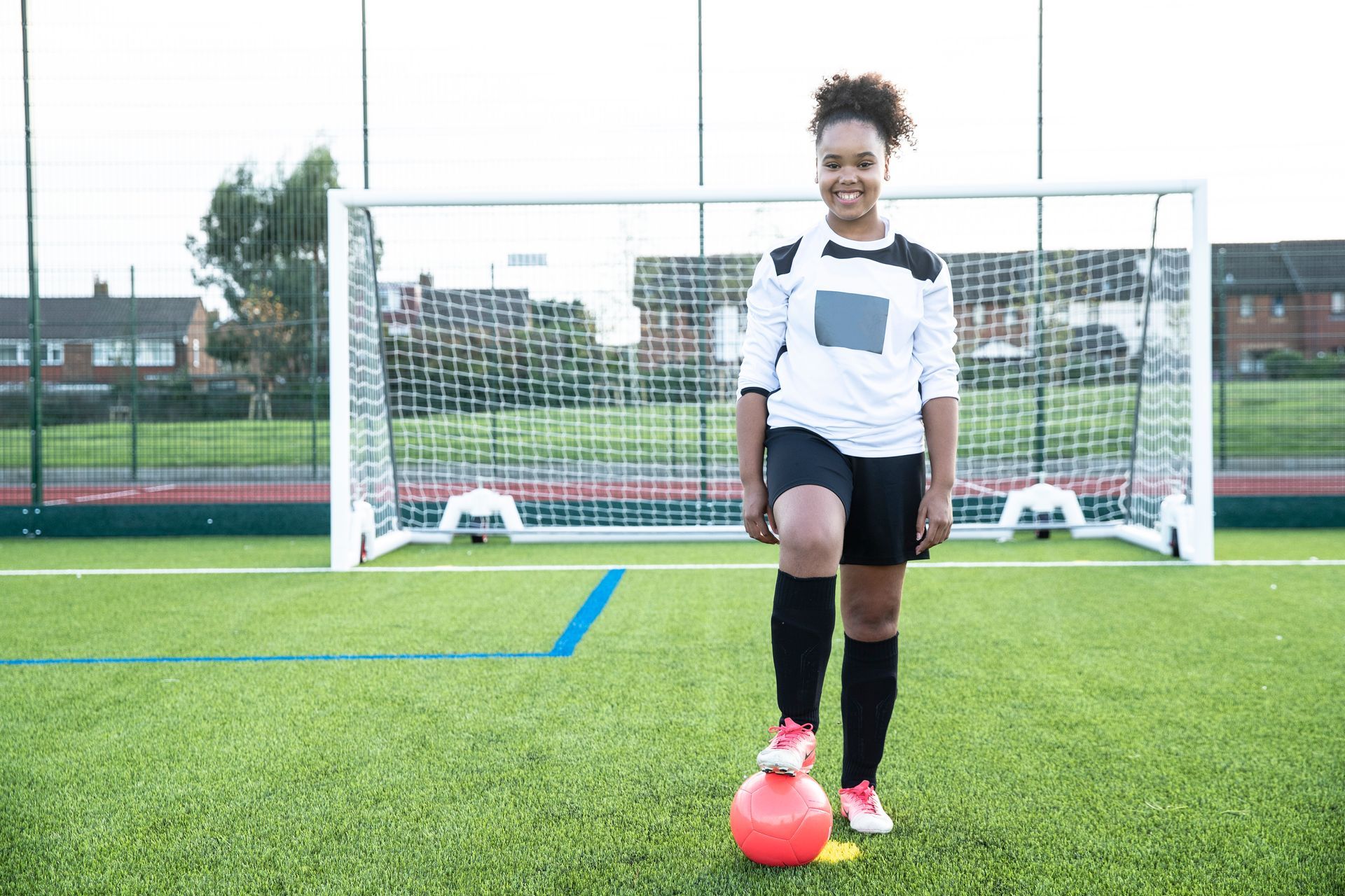 Soccer player in white and black uniform smiles on a green field, ball at her foot in front of a goal.