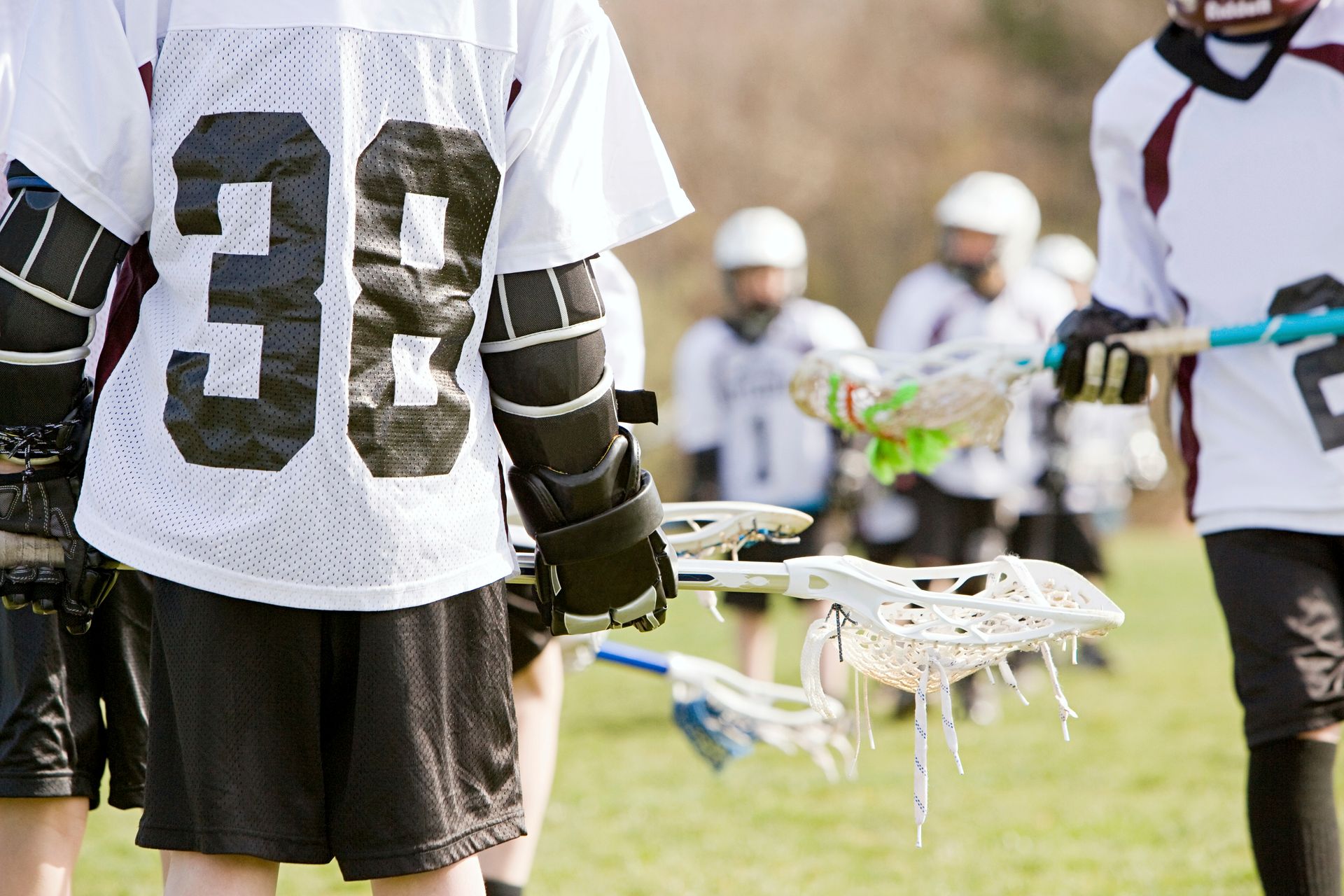 Lacrosse players on a grassy field, wearing white jerseys with black numbers, holding lacrosse sticks.