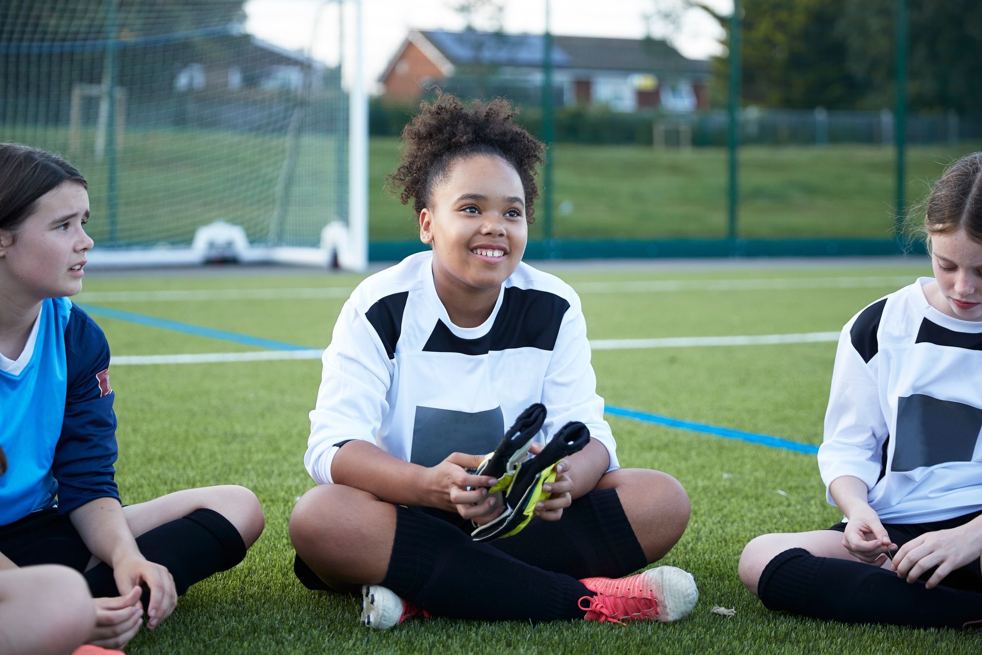 Three soccer players sit on a green field. Girl in center holds cleats, smiling. Others look on.
