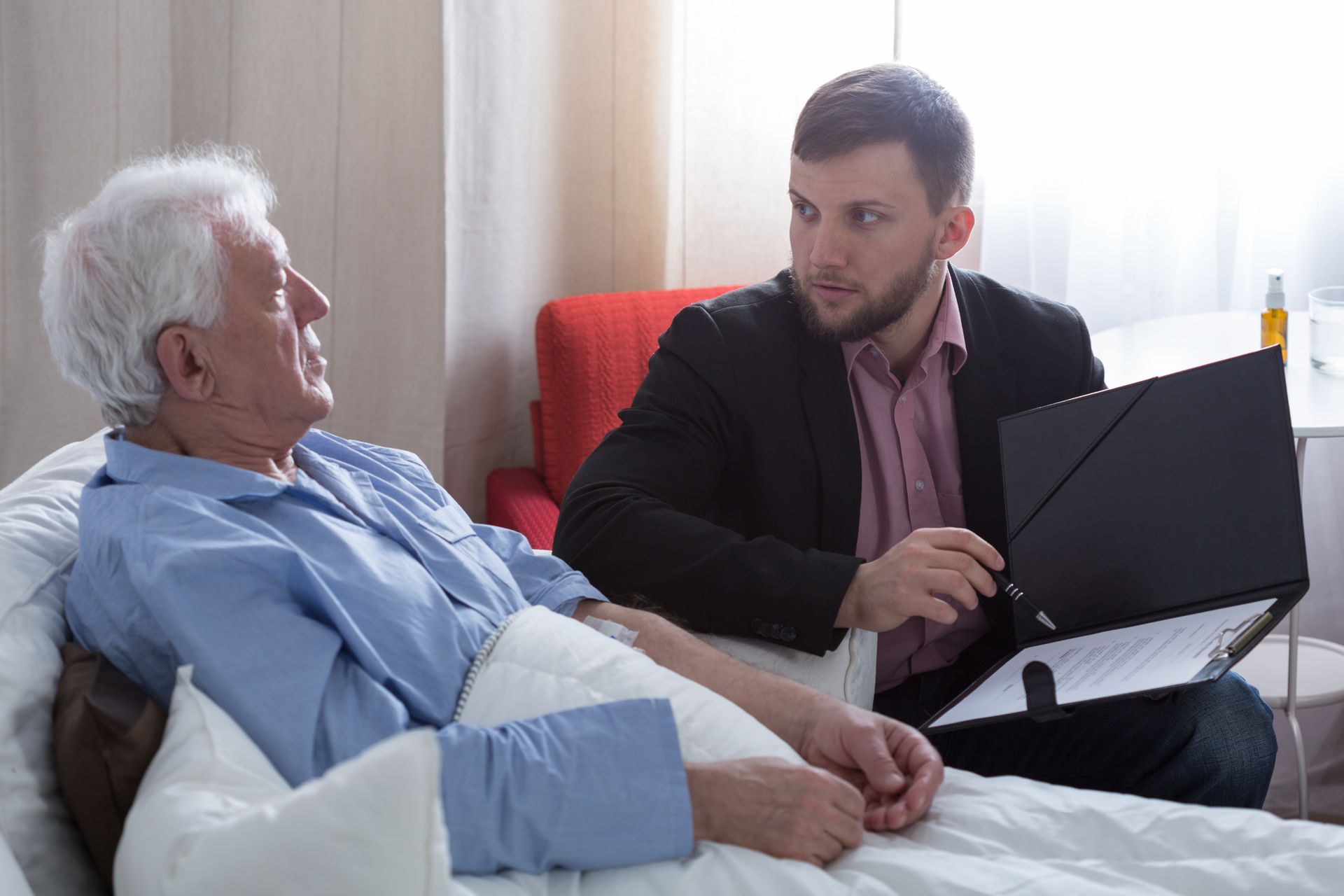 Caregiver talks with a patient in bed while holding a laptop in a bright room.