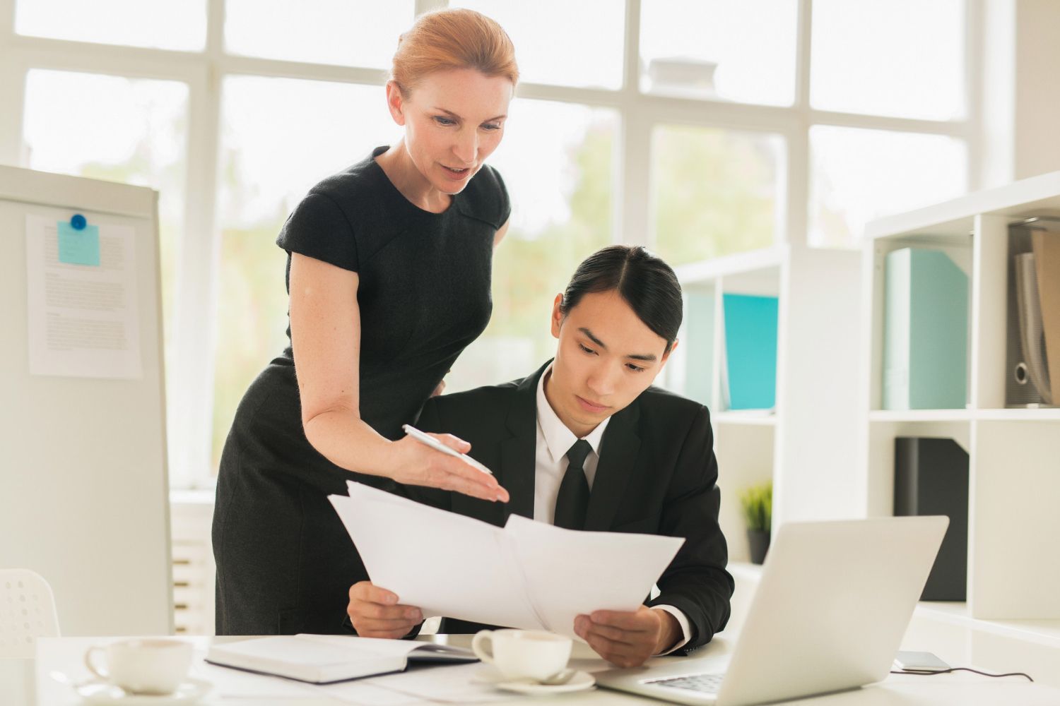 Woman in black dress pointing at papers held by a man in a suit at a desk, looking at a laptop.