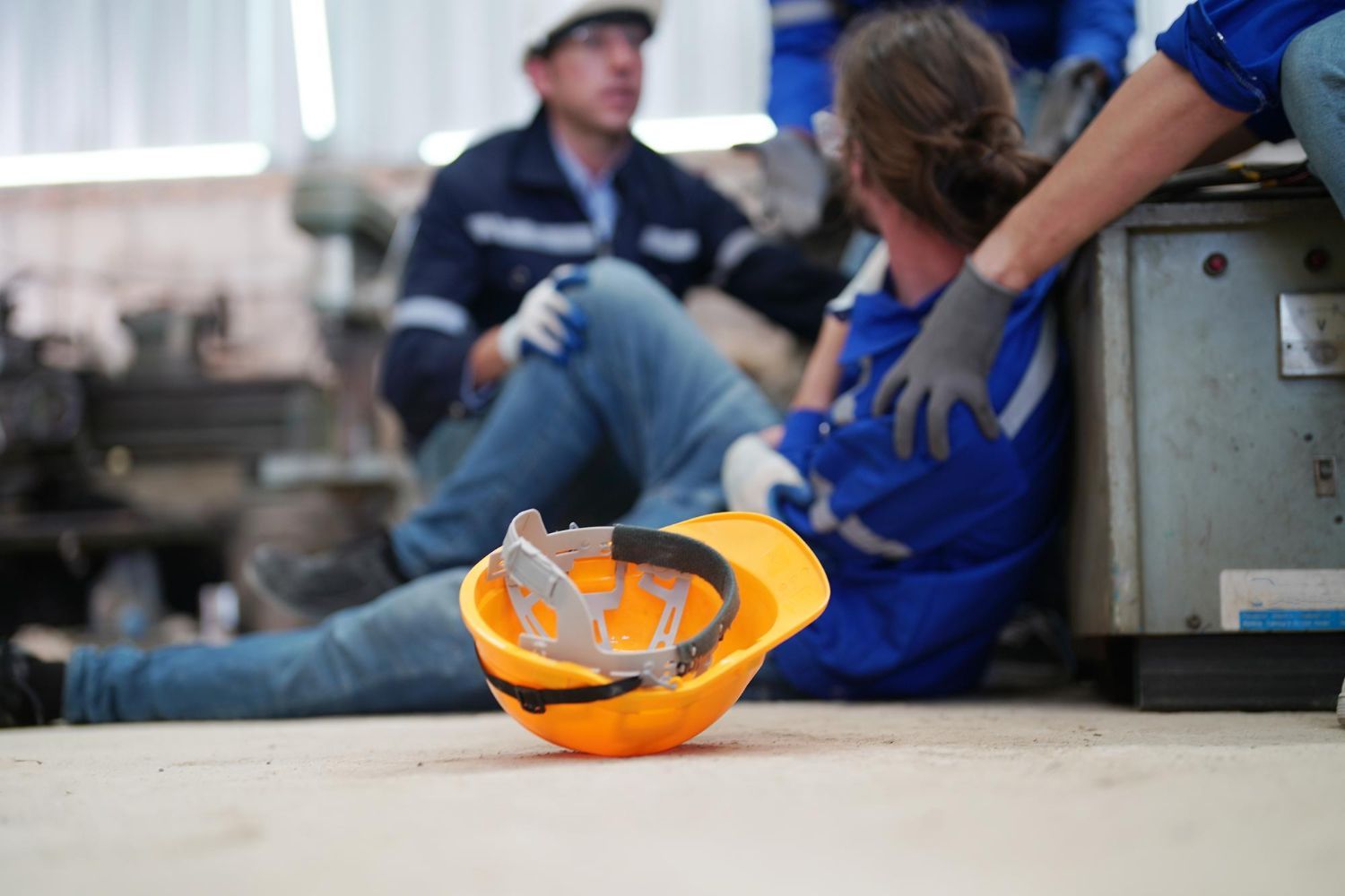 A worker injured in a factory, with colleagues assisting. Yellow hard hat on floor.
