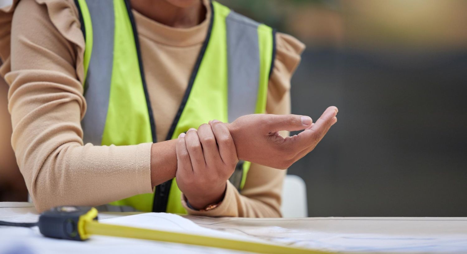 Person in safety vest holding their wrist, possibly in pain, with a measuring tape visible.
