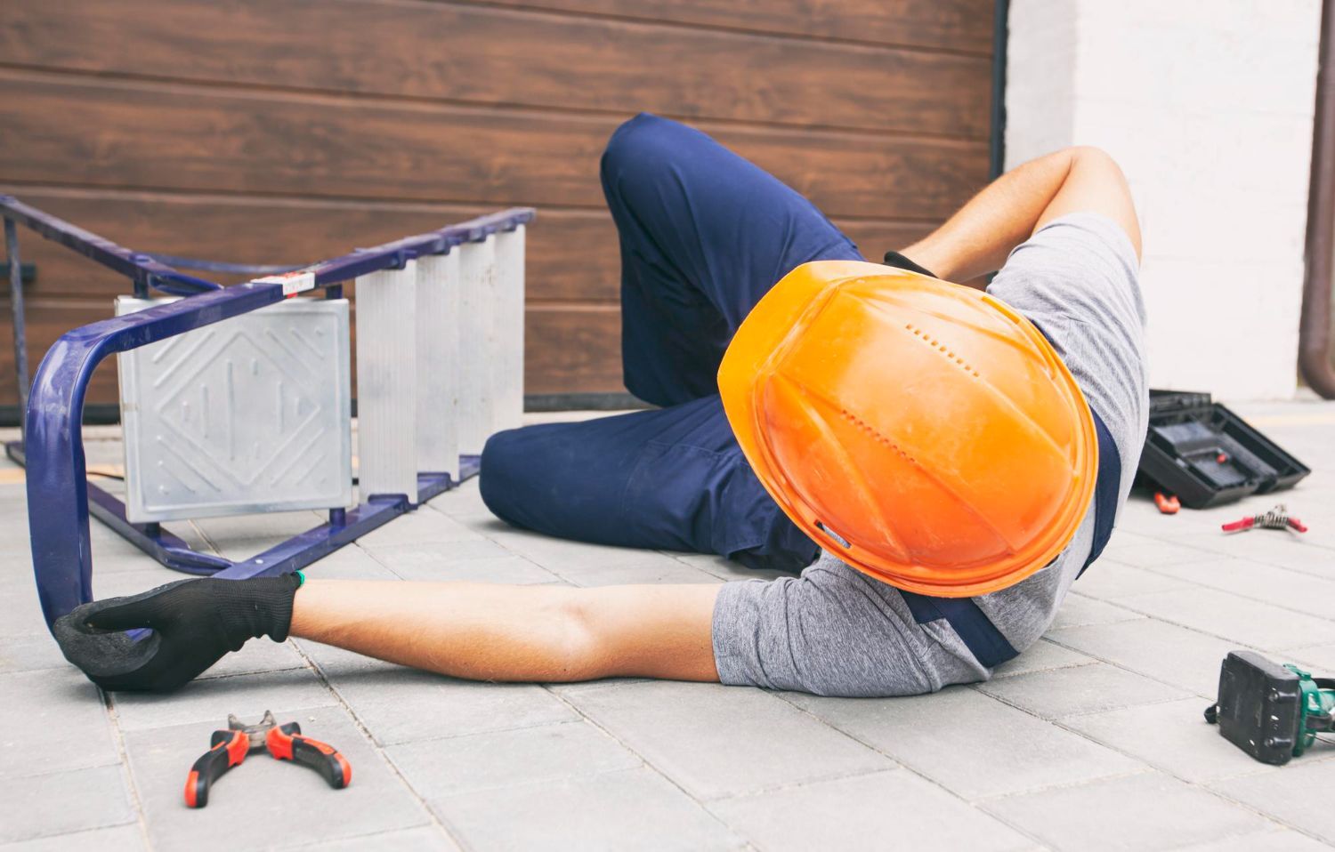 Person in hard hat lies on ground next to fallen ladder and tools.
