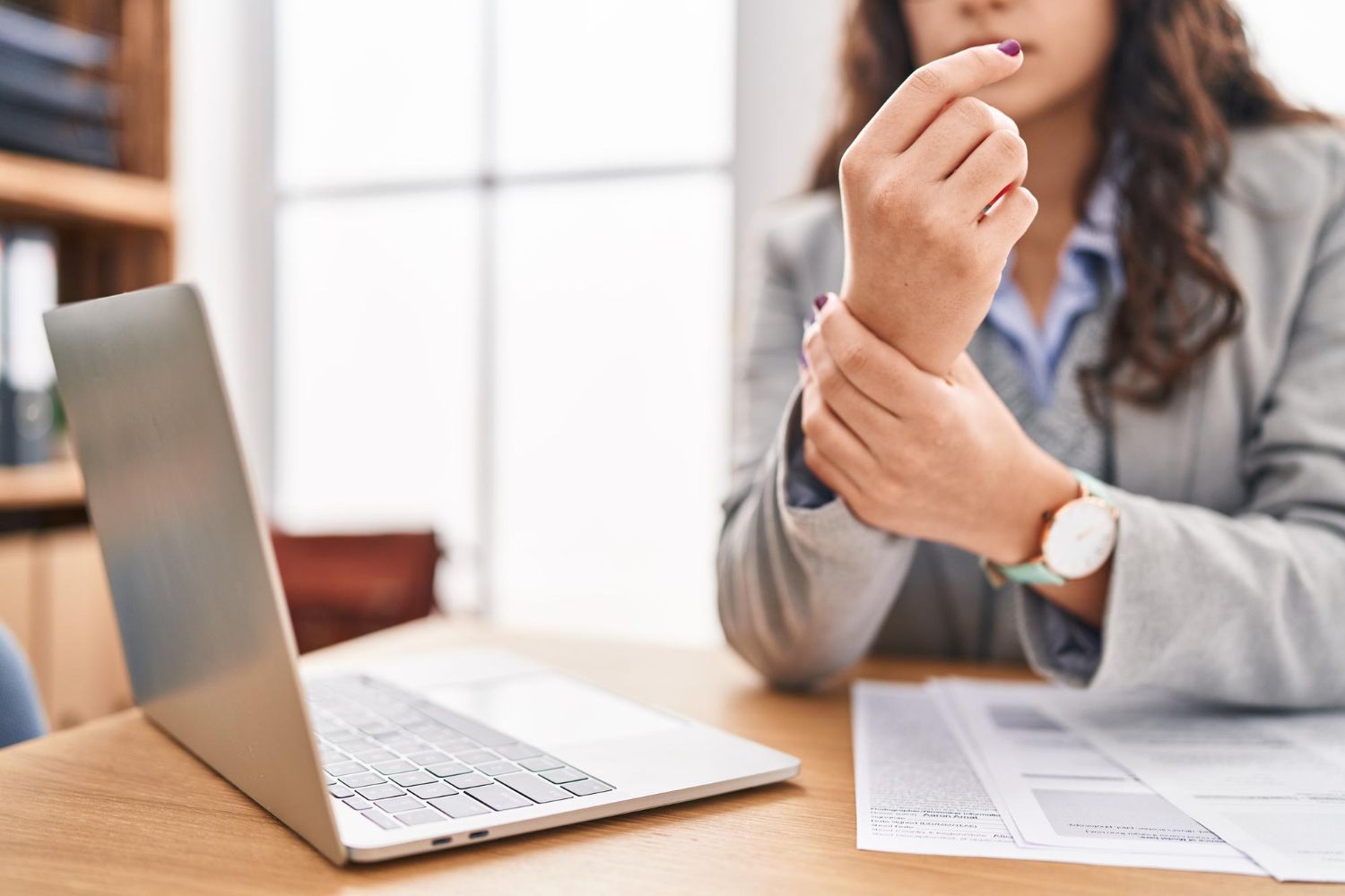 Woman at a desk with a laptop, touching her wrist, possibly experiencing pain.
