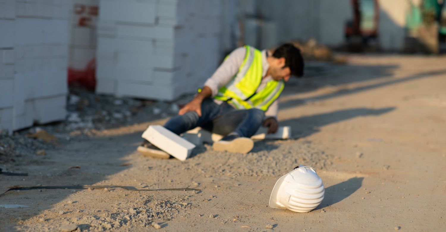 Construction worker on ground after fall, wearing a yellow safety vest; hard hat on the ground.