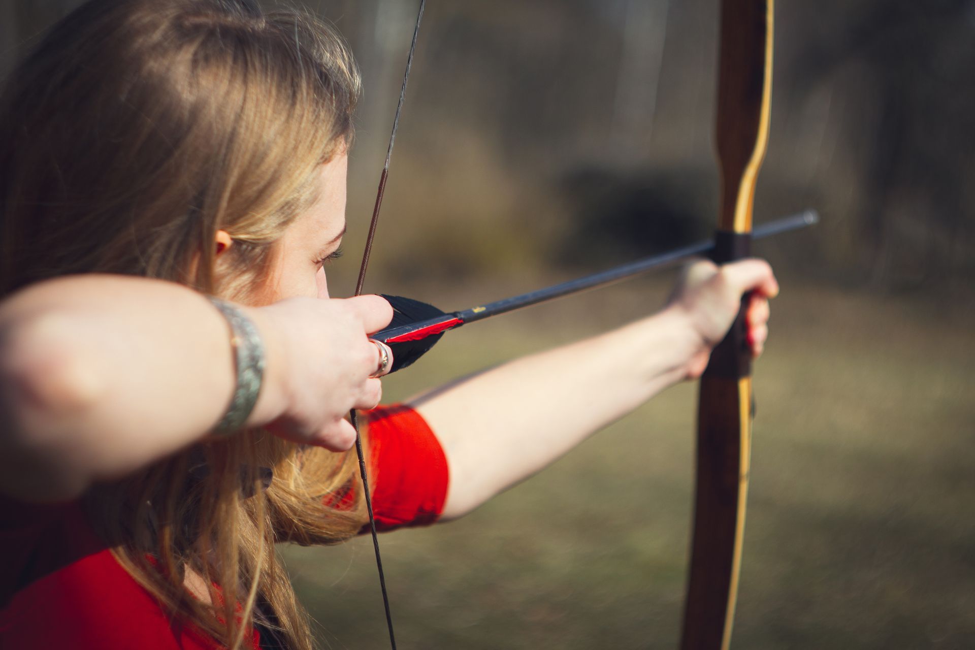 Woman in red shirt drawing back a bow and arrow, aiming towards a target. Outdoor setting.