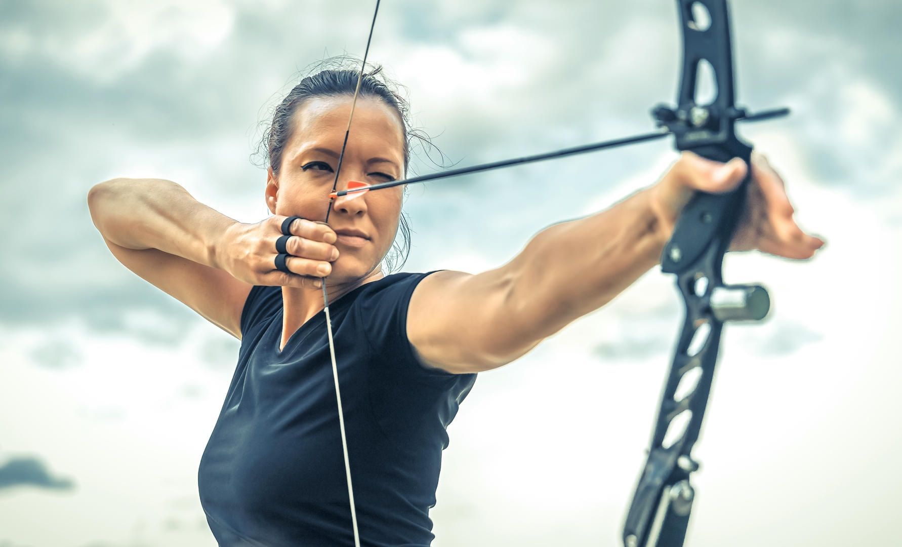 Woman with black compound bow, drawing the string back, outdoors.