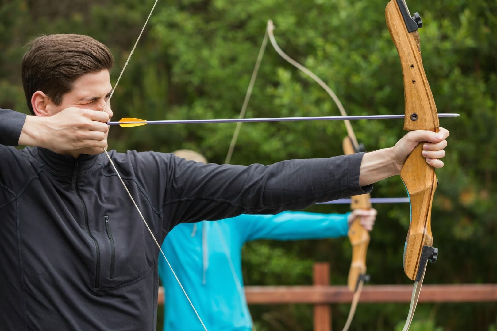 Man aiming a bow and arrow at a target. He's outdoors, with a focused expression. Another archer is in the background.