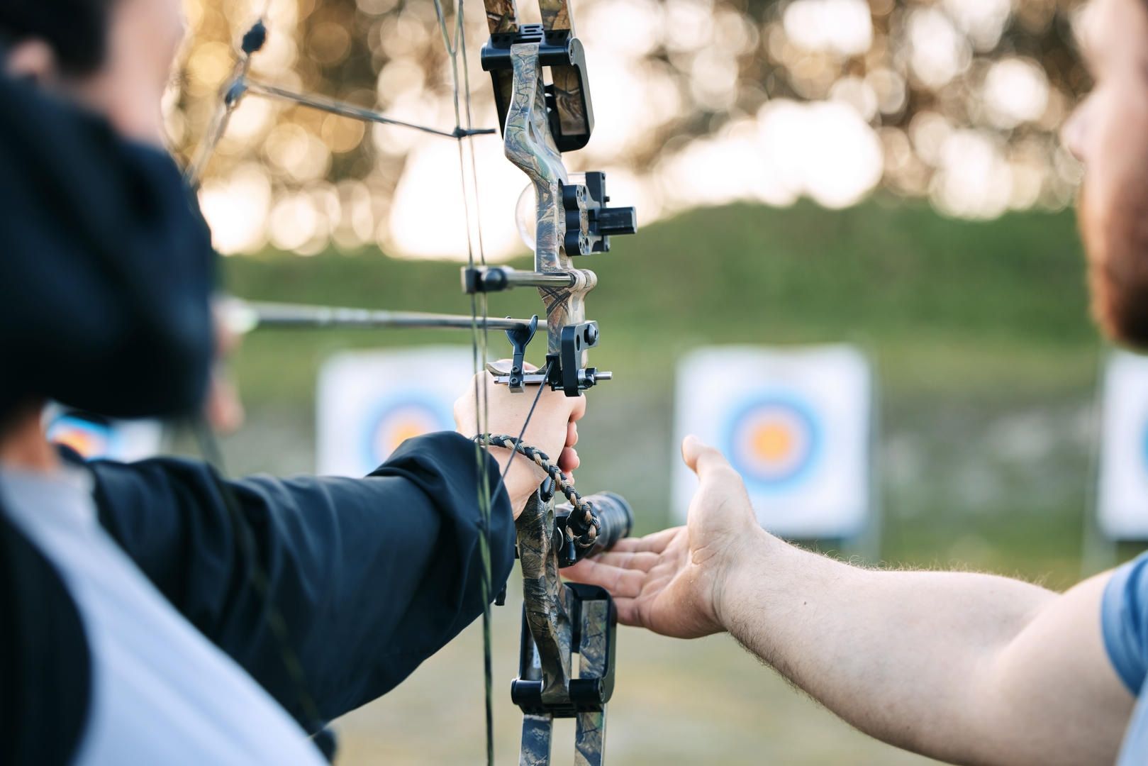 Two people practicing archery; one drawing back bow, other guiding. Targets in background.