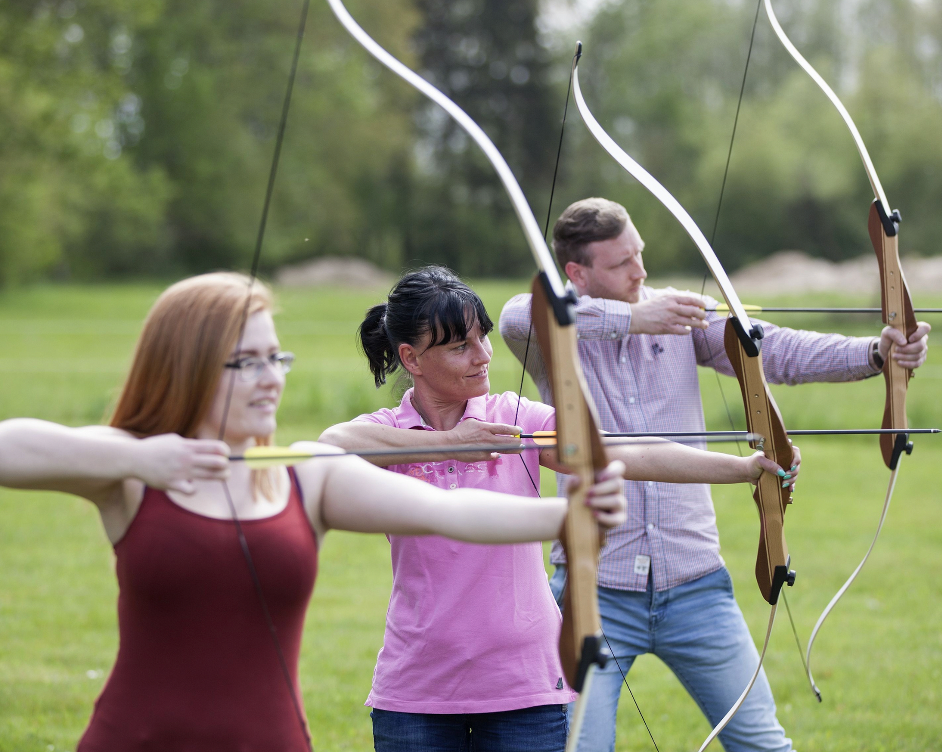 Three people aiming bows in a grassy field. Two are focused; one smiles.