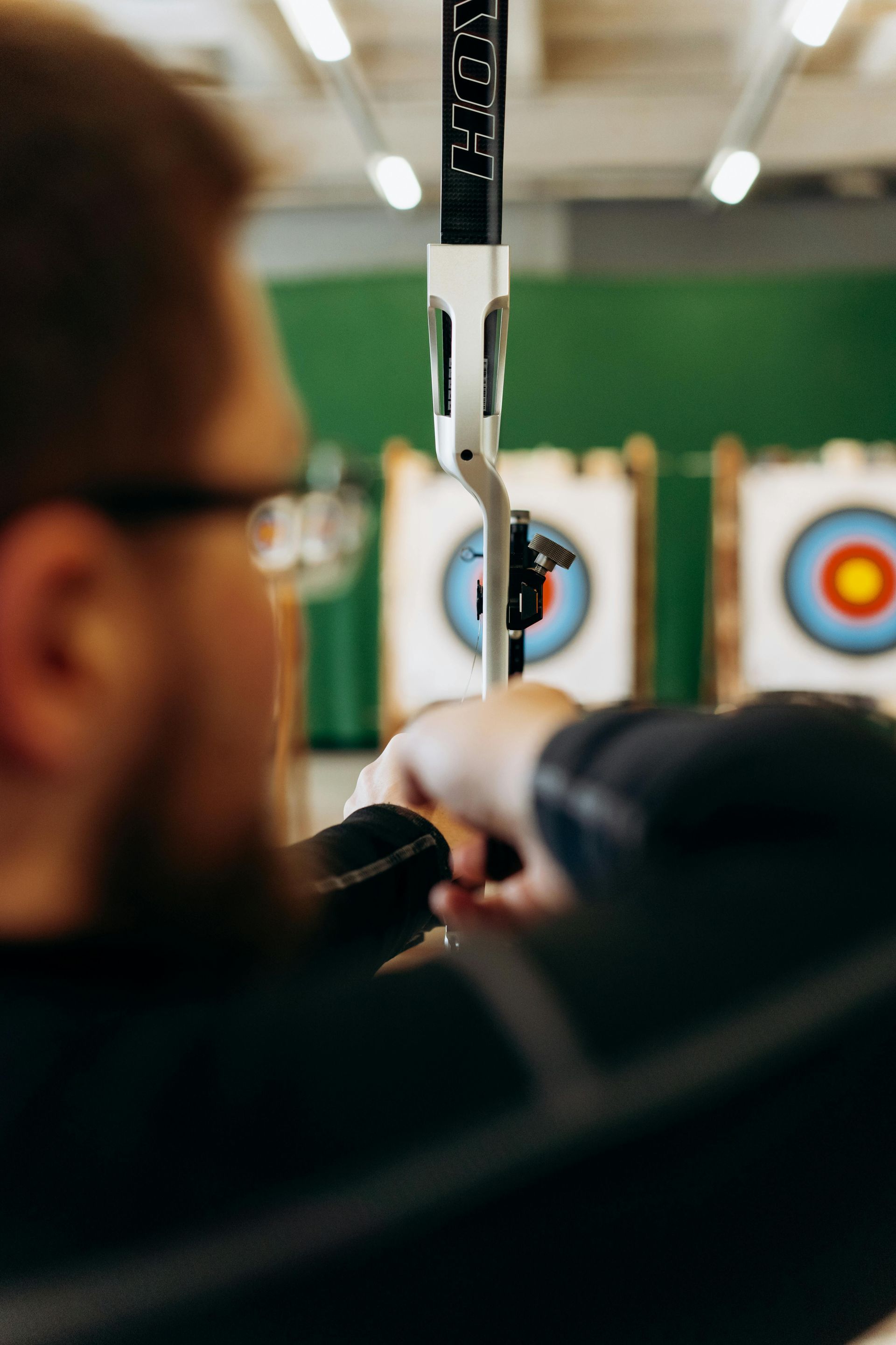 Archer aiming at target in an indoor range; targets visible in the background, green wall.