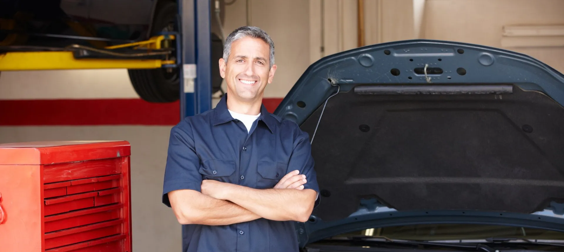 A man is standing in front of a car with the hood open in a garage.