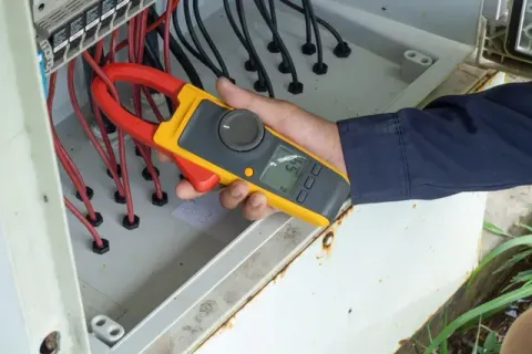 Hand holding a yellow digital multimeter testing wires in an electrical control box