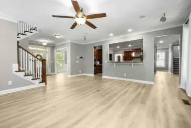 Spacious open-concept living room with light wood floors, gray walls, staircase, ceiling fan, and kitchen pass-through.