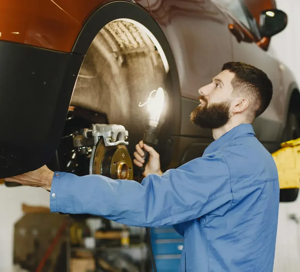 Mechanic in blue coveralls examines car brakes, using a light, in a garage.