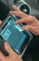 Person using a diagnostic tool connected to a car. Tool displays a screen, inside the vehicle.