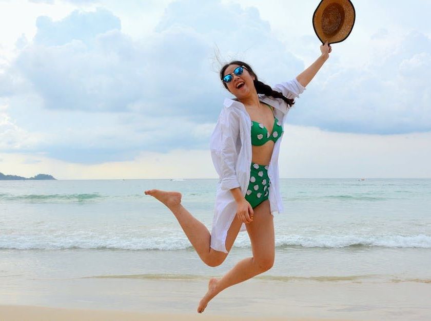 Woman in green polka dot bikini jumping joyfully on a beach, holding a straw hat, smiling.