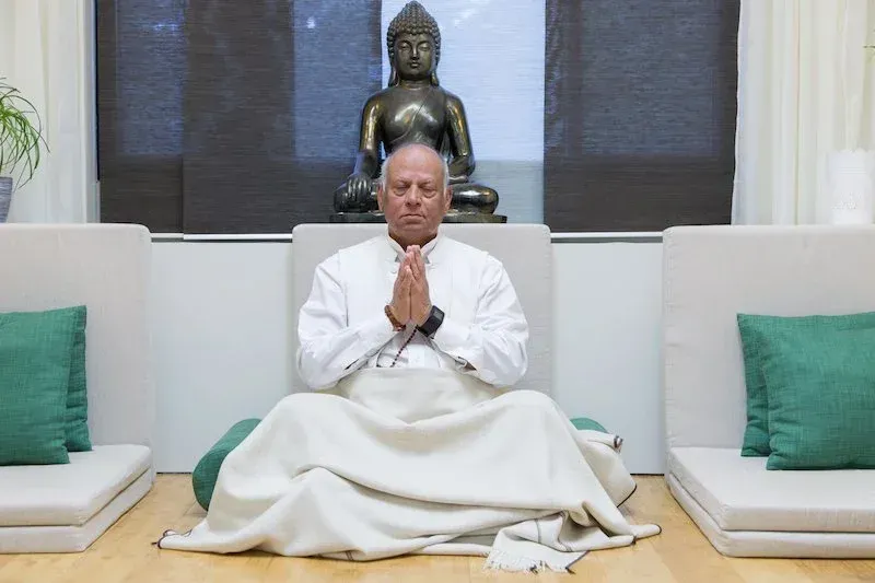 Man meditating in front of Buddha statue; hands clasped, white robe, green cushions.