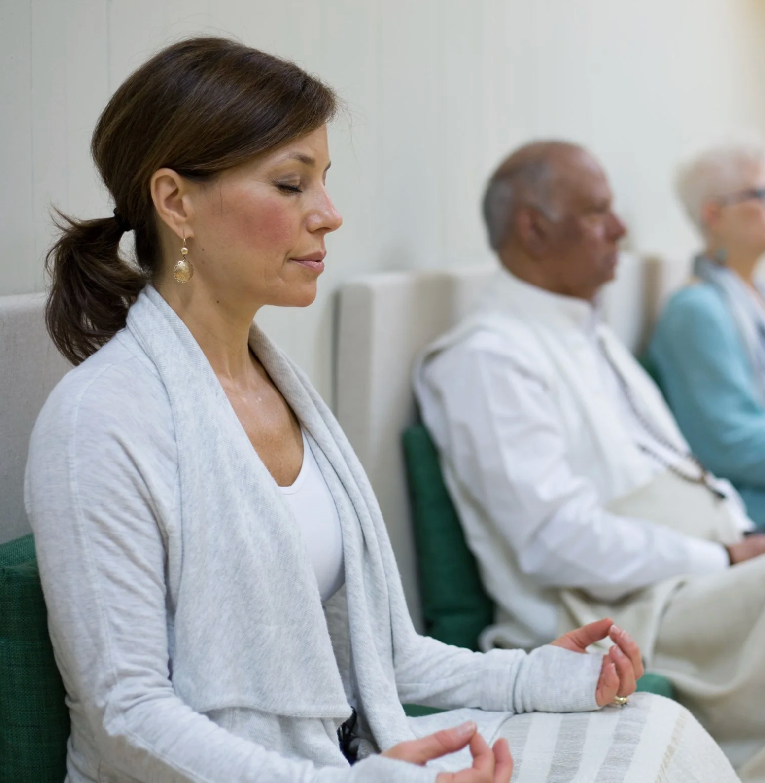 Woman meditating with eyes closed, seated indoors next to others; serene expressions.