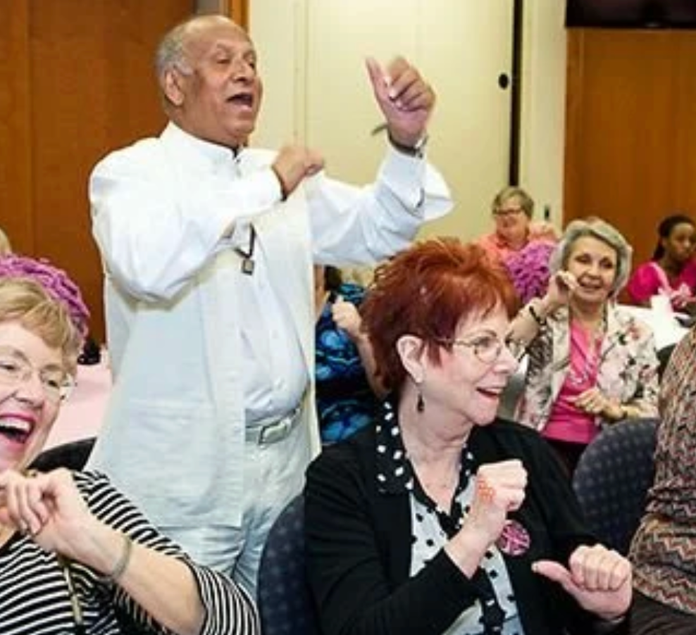 Man dancing, leading a group in a room. People in attendance smile and move to the music.