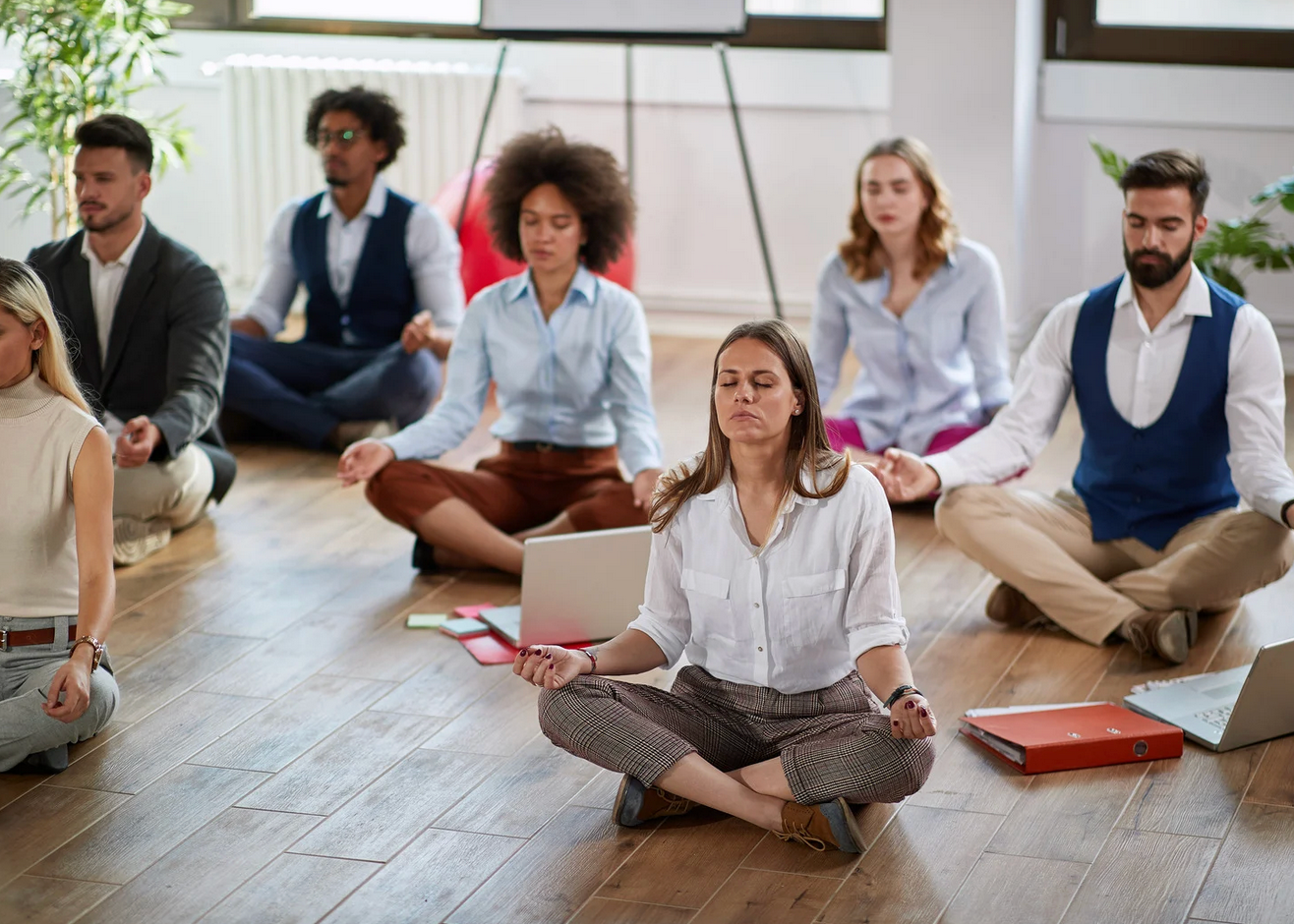 People in business attire meditating on a wooden floor in an office.