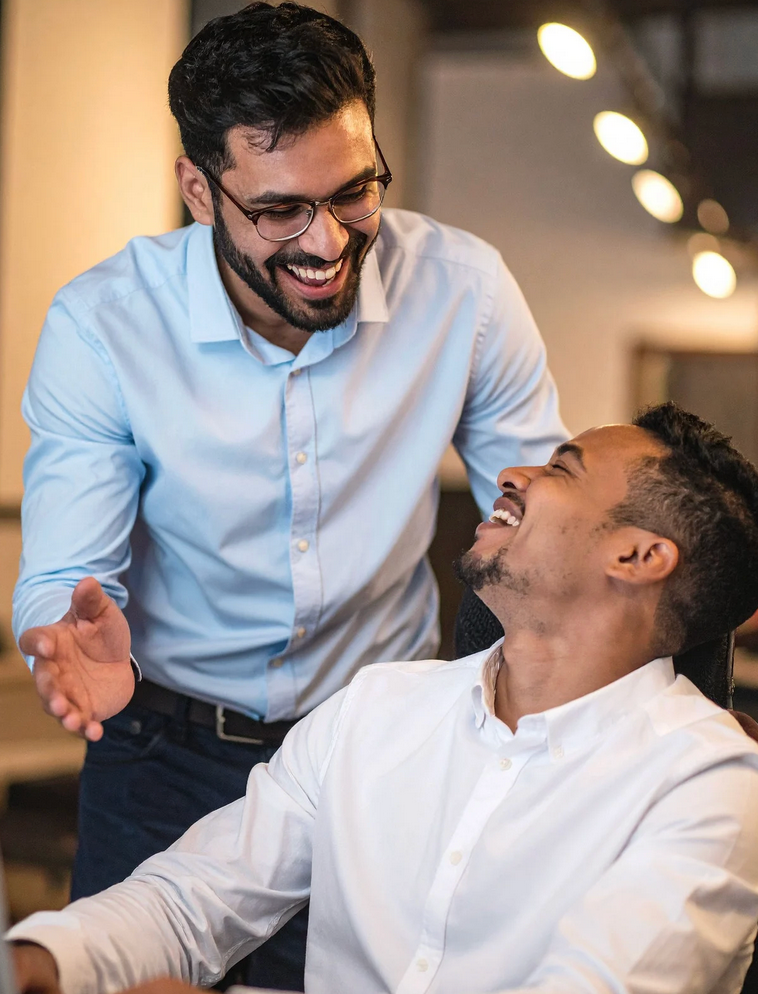 Two men in office setting laughing; one wears glasses and light blue shirt, the other wears white shirt.