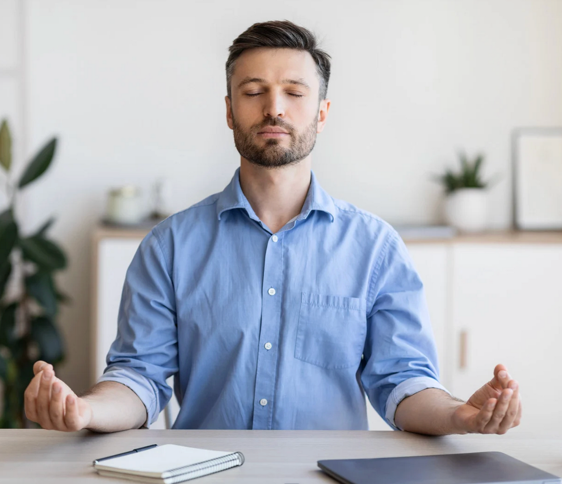 Man with eyes closed, meditating at a desk; hands up, office setting.