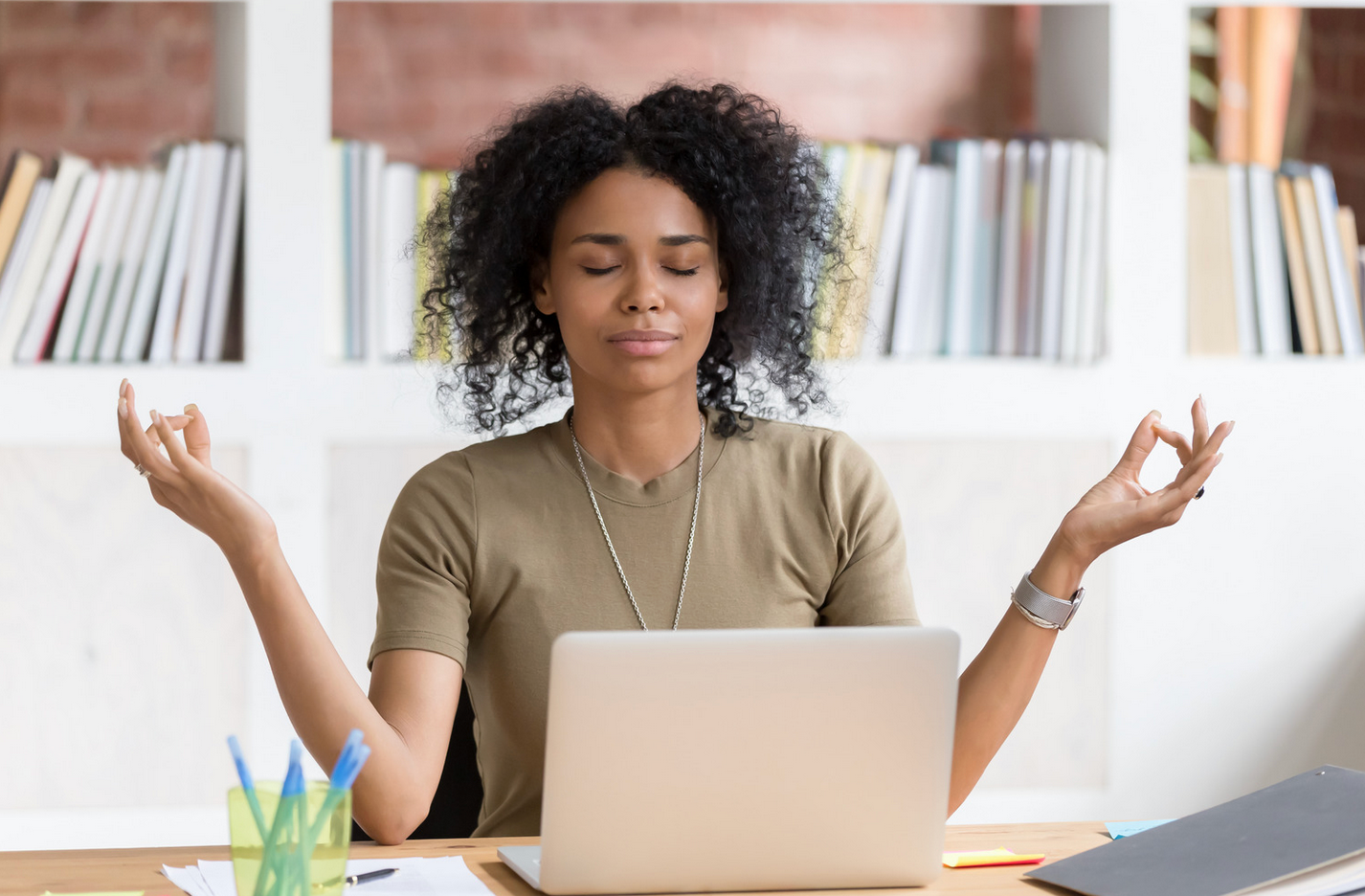 Woman meditating at a desk, eyes closed, hands in mudra pose, laptop in front, books in the background.