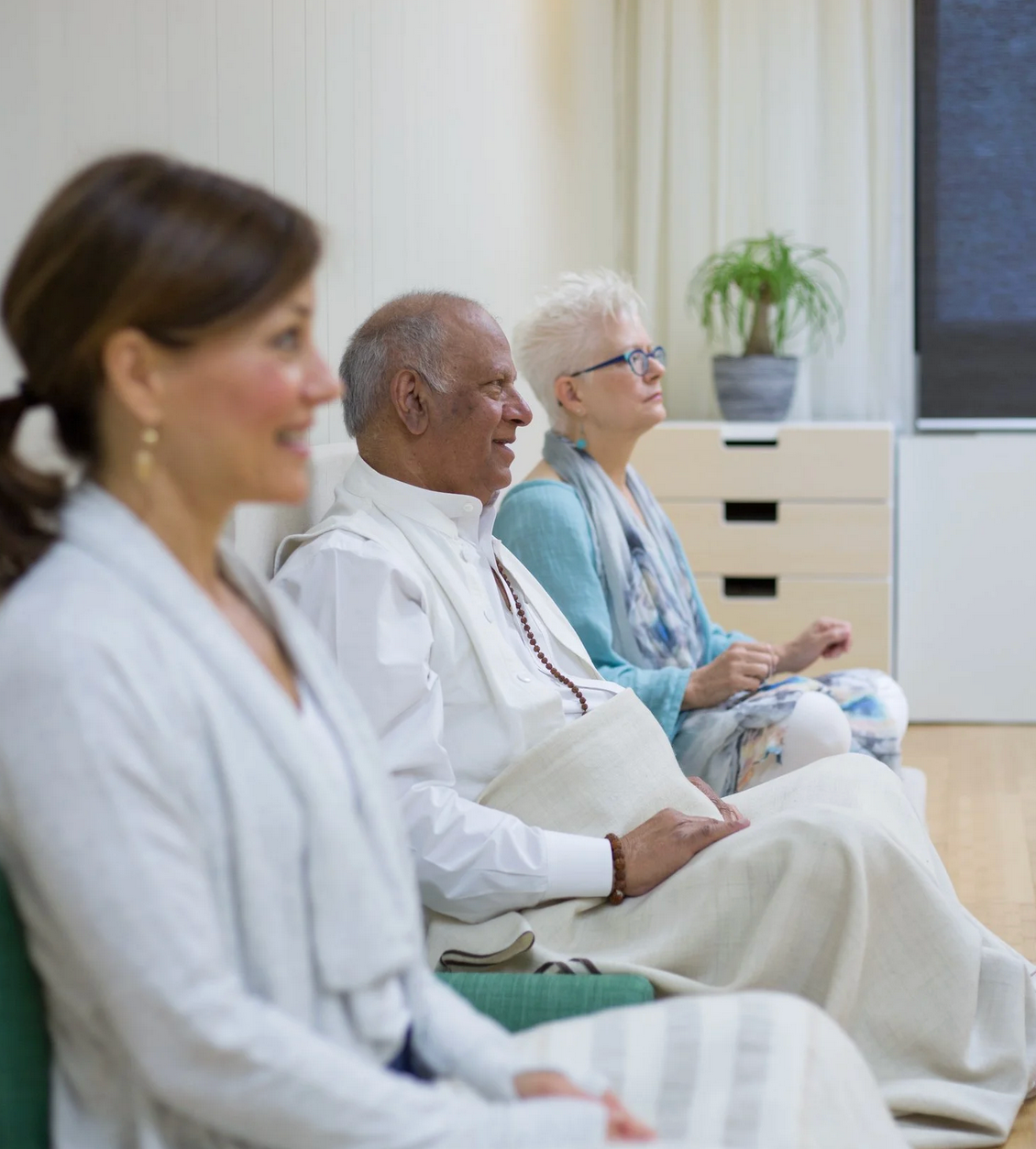 People seated and meditating in a light-filled room; a woman and two others with eyes closed.