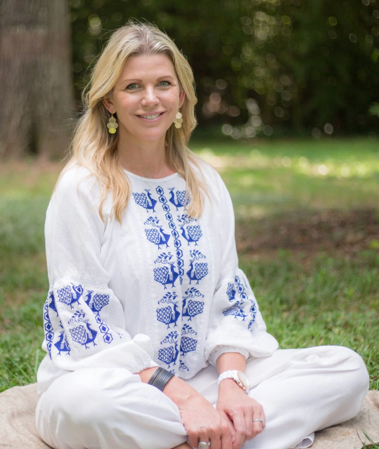 Woman in embroidered white shirt and pants sits outdoors, smiling.