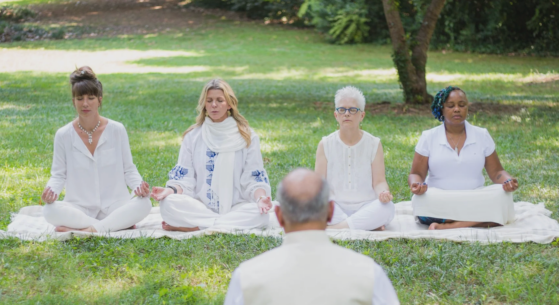 Four people in white clothing meditate on a mat outdoors, guided by a person facing them.
