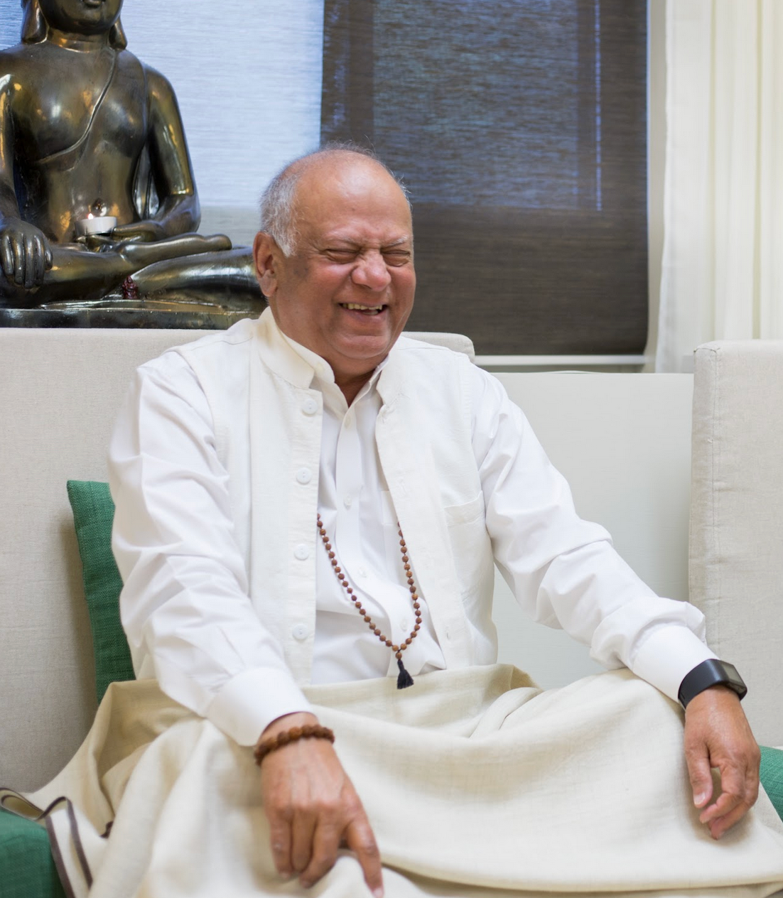 Man in white smiling, sitting, with mala beads. Buddha statue in background.