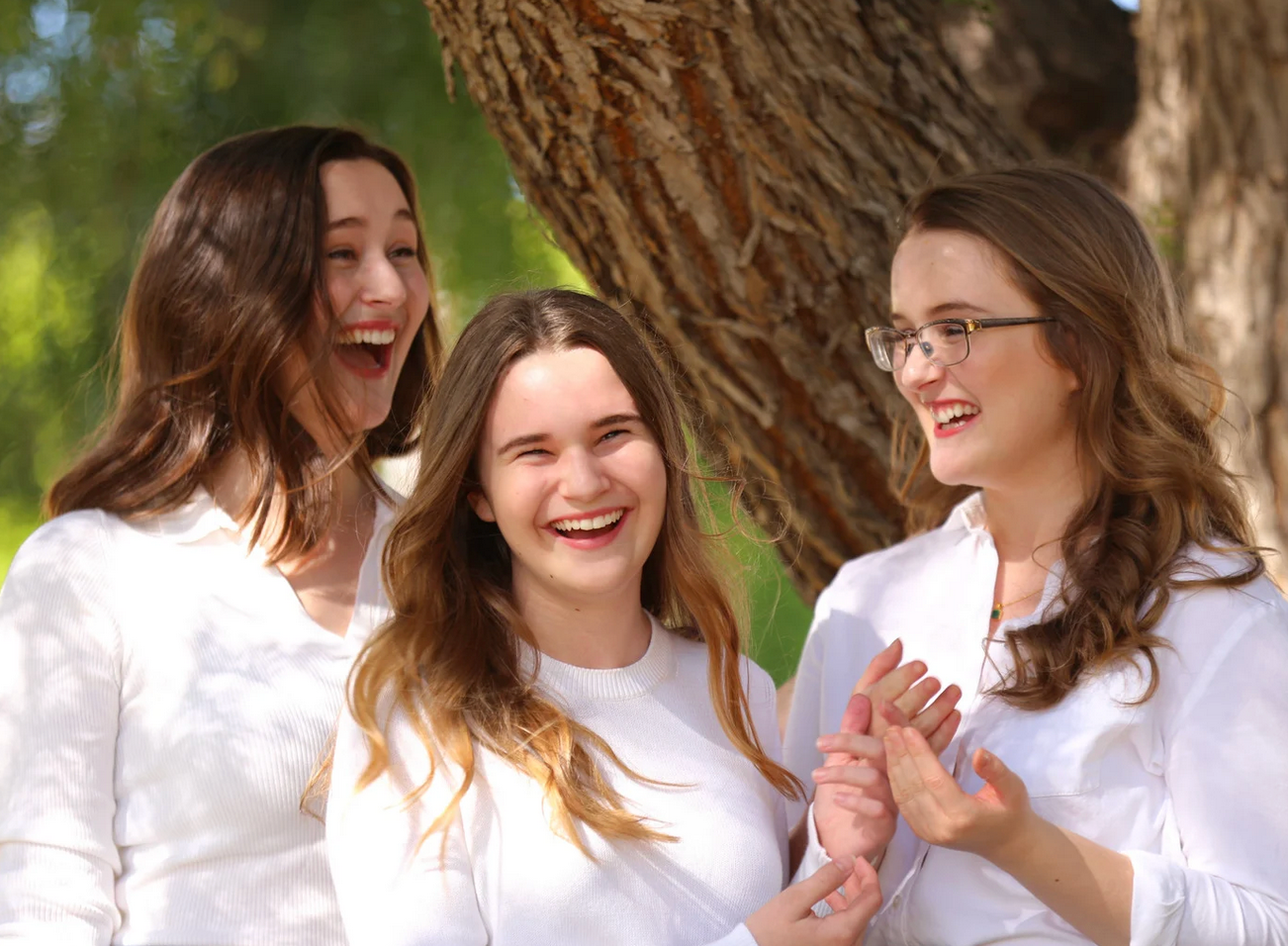 Three women in white laughing outdoors by a tree.