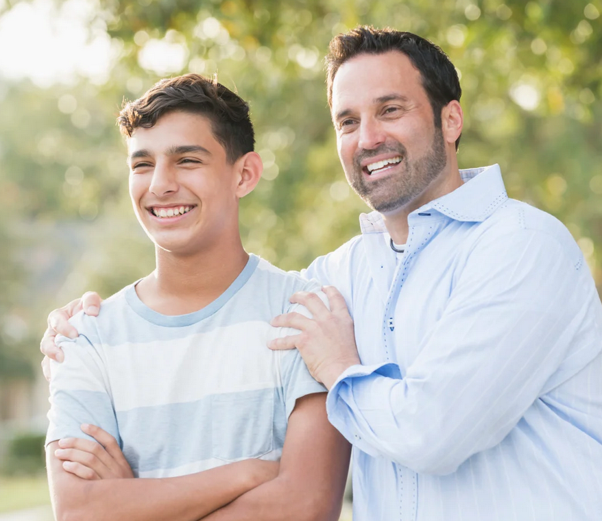 Woman with long gray hair, wearing a brown shawl, smiles, hand on chest; outdoor setting.