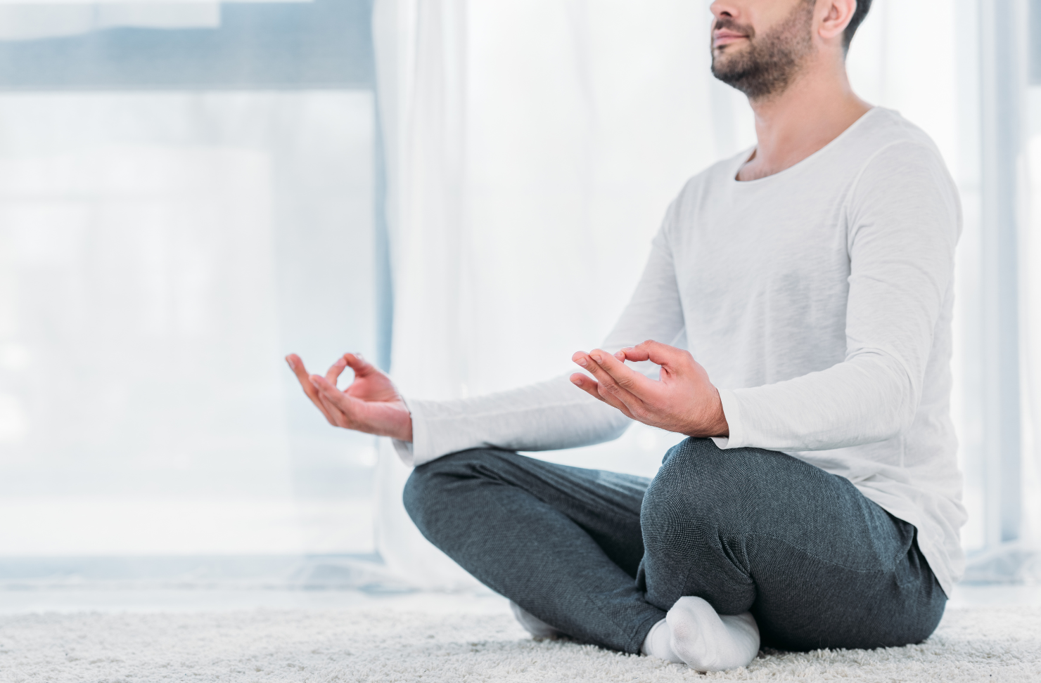 Man in white shirt and gray pants meditating indoors, seated on the floor in lotus position.