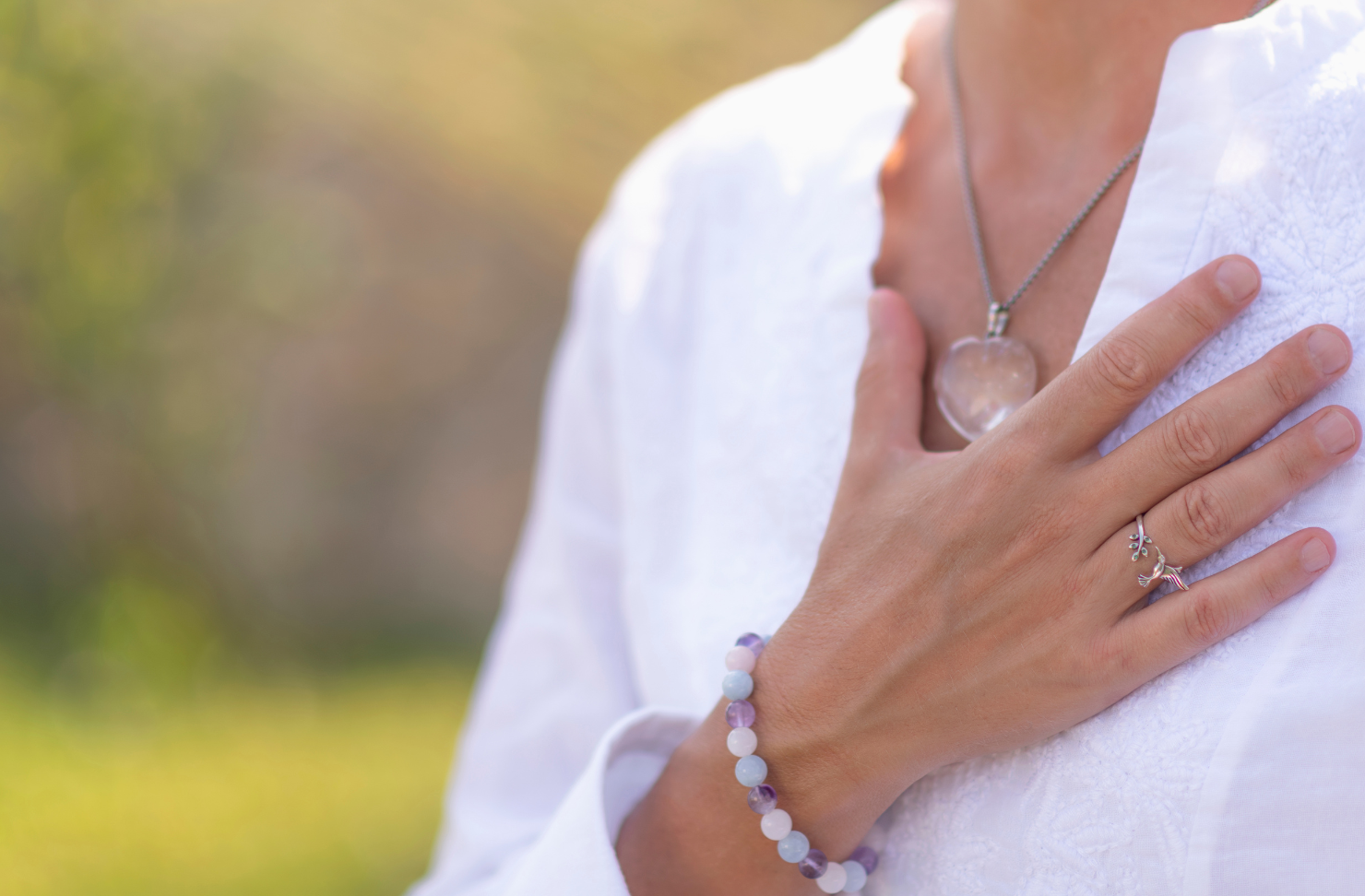 Woman with hand over chest, wearing white, outdoors, with necklace and bracelet.