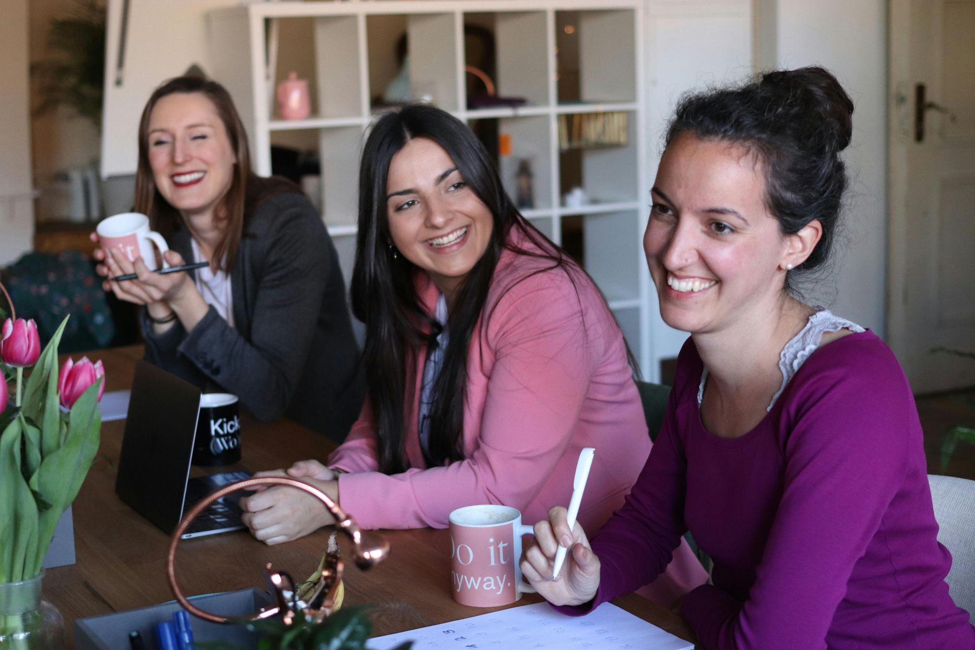 Three women smiling around a table. One holds a mug, another a pen, with tulips nearby. Bright room setting.