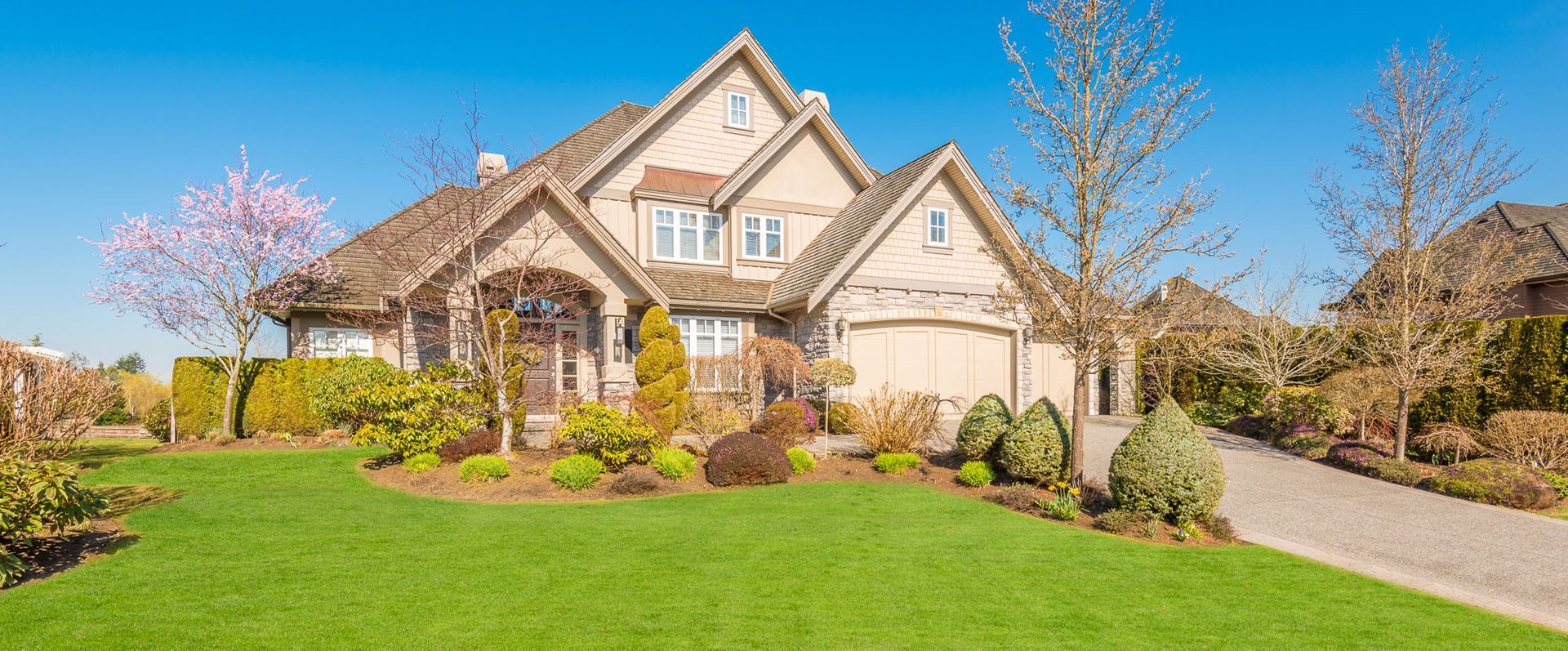 Large suburban house with beige siding, manicured lawn, trees, and a curved driveway on a sunny day