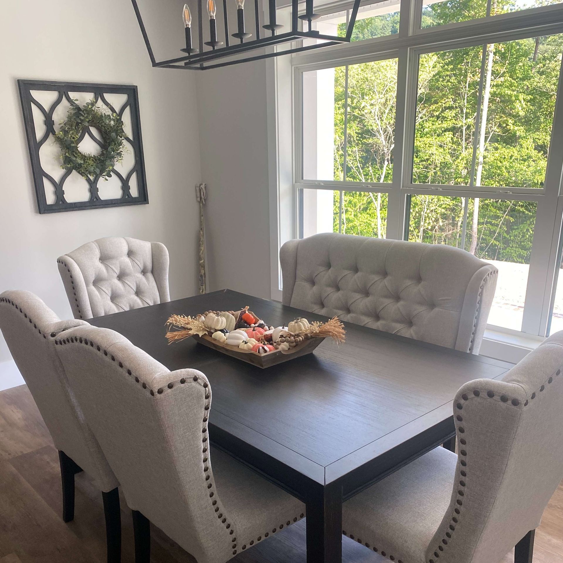 Dining room with tufted gray chairs around a dark table by a bright window and wall wreath decor