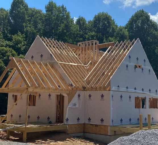 Partially built house with exposed wooden roof framing and white house wrap under a blue sky