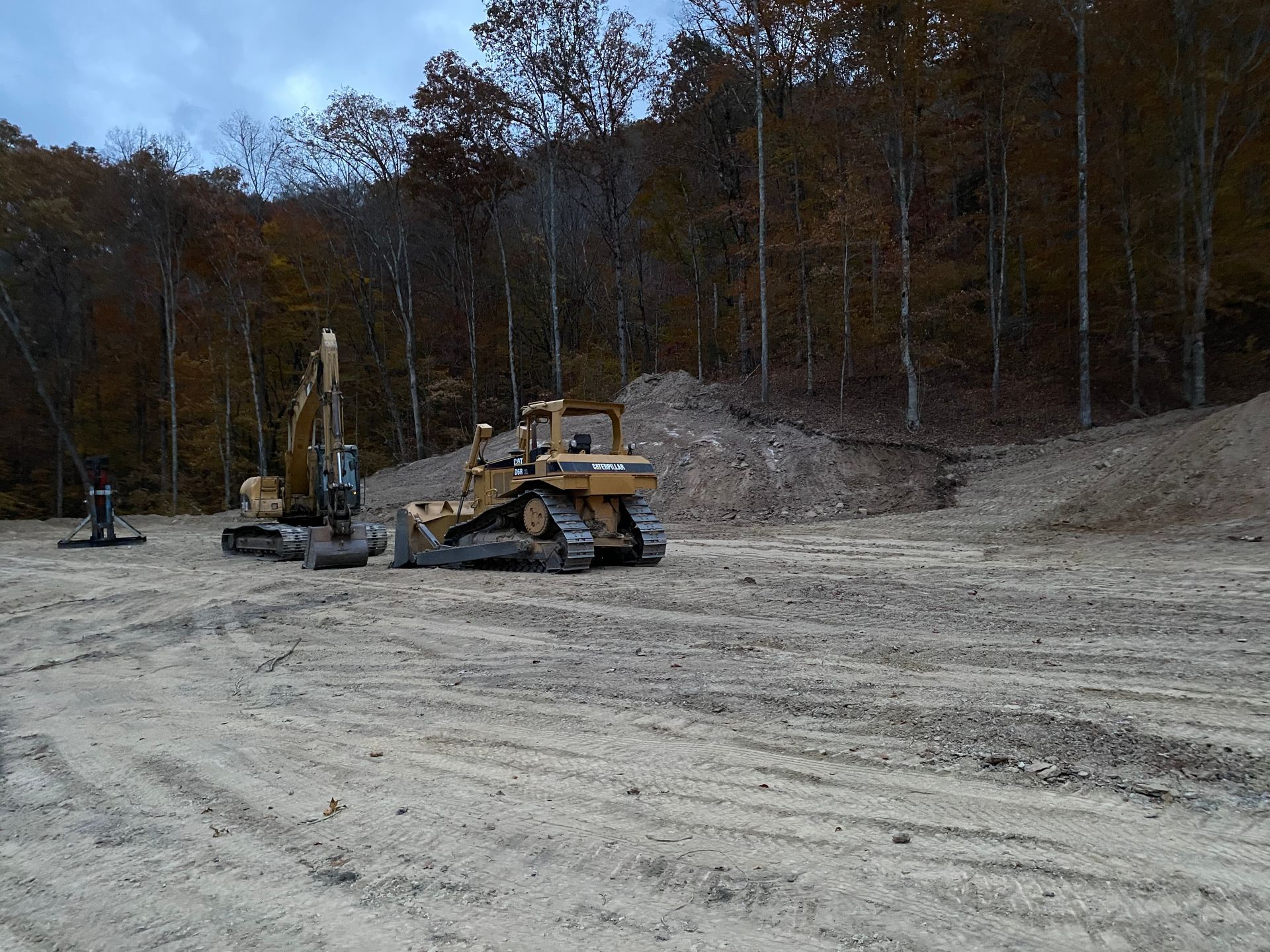 Yellow road grader on a dirt lot with a wooded hillside in the background