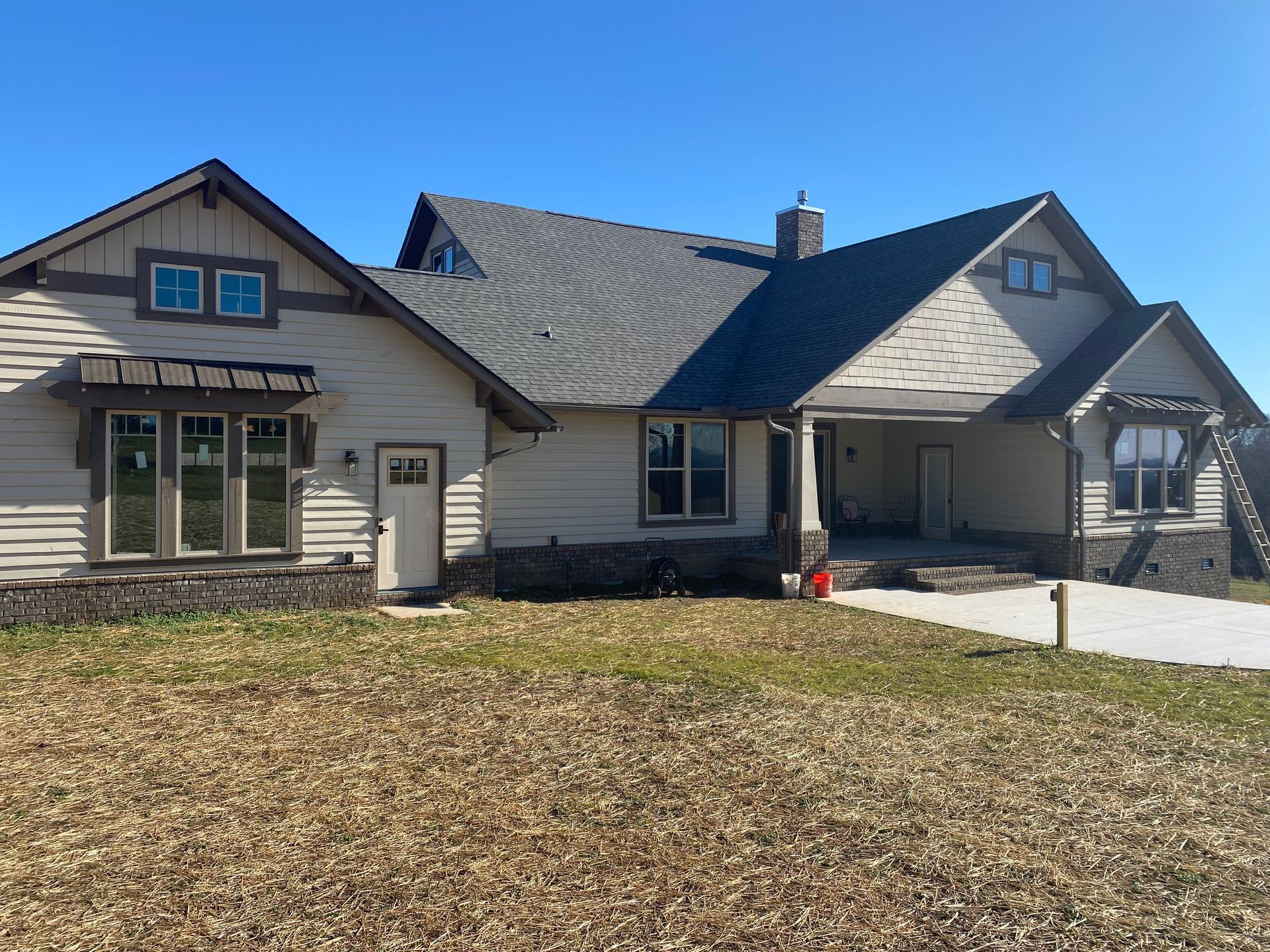 Single-story beige house with dark roof, front porch, and a bare lawn under a clear blue sky